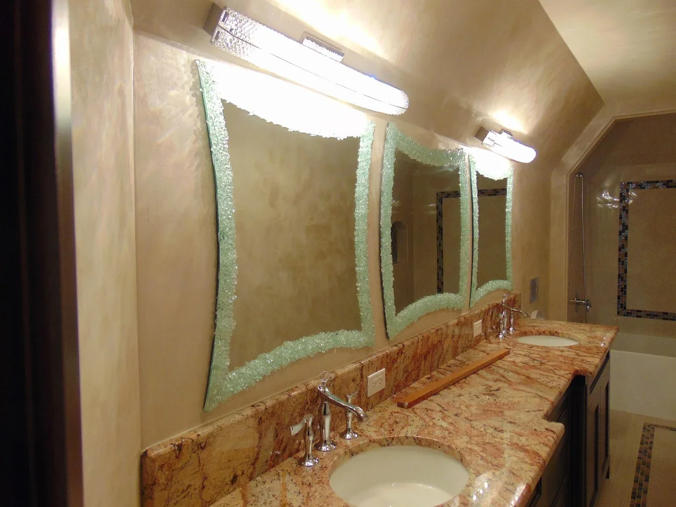 Bathroom with a double vanity, granite countertop, two sinks, and two broken mirrors bordered with decorative mirror frames. There are two wall-mounted lights above each mirror, a bathtub visible in the background, and beige walls.