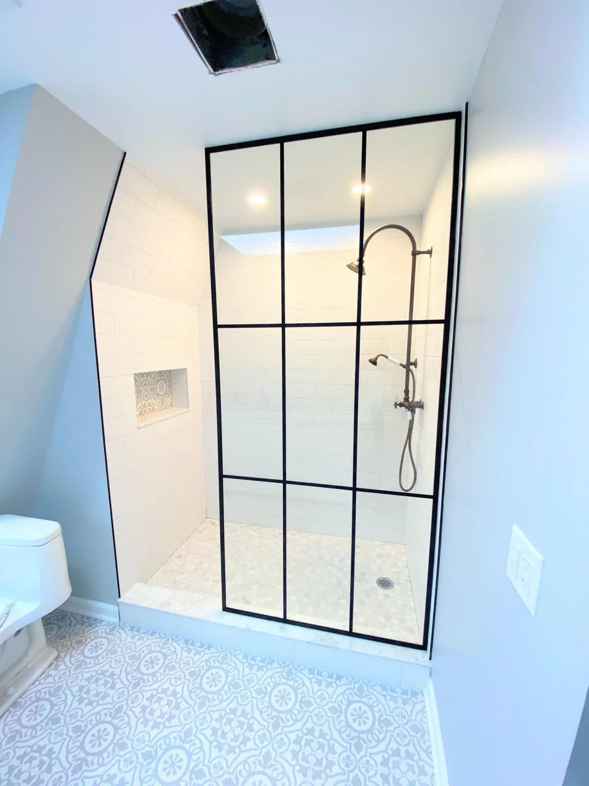 Modern walk-in shower with black metal-framed glass enclosure, white tile walls, and pebble tile floor. A niche with patterned tiles is built into the wall. The ceiling has a skylight and a showerhead on the right wall.