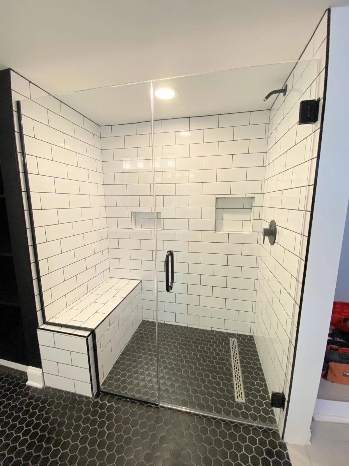 Modern walk-in shower with white subway tile walls, black grout, a built-in bench, and hexagonal black floor tiles. It has a glass door and niche shelves for toiletries.