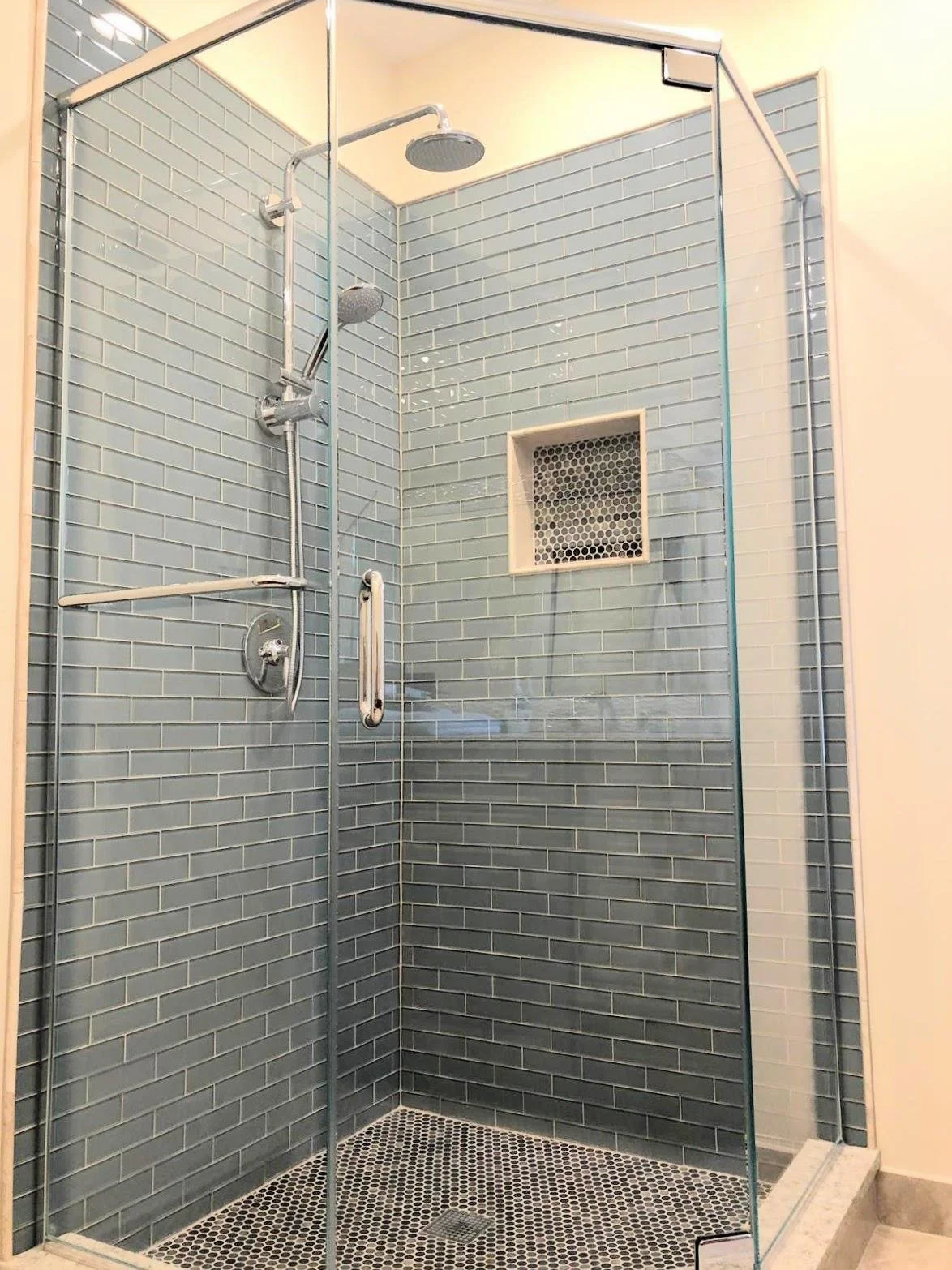 Glass-enclosed shower with gray subway tiles, a rain showerhead, a handheld shower, a small niche, and a black and white mosaic tile floor.