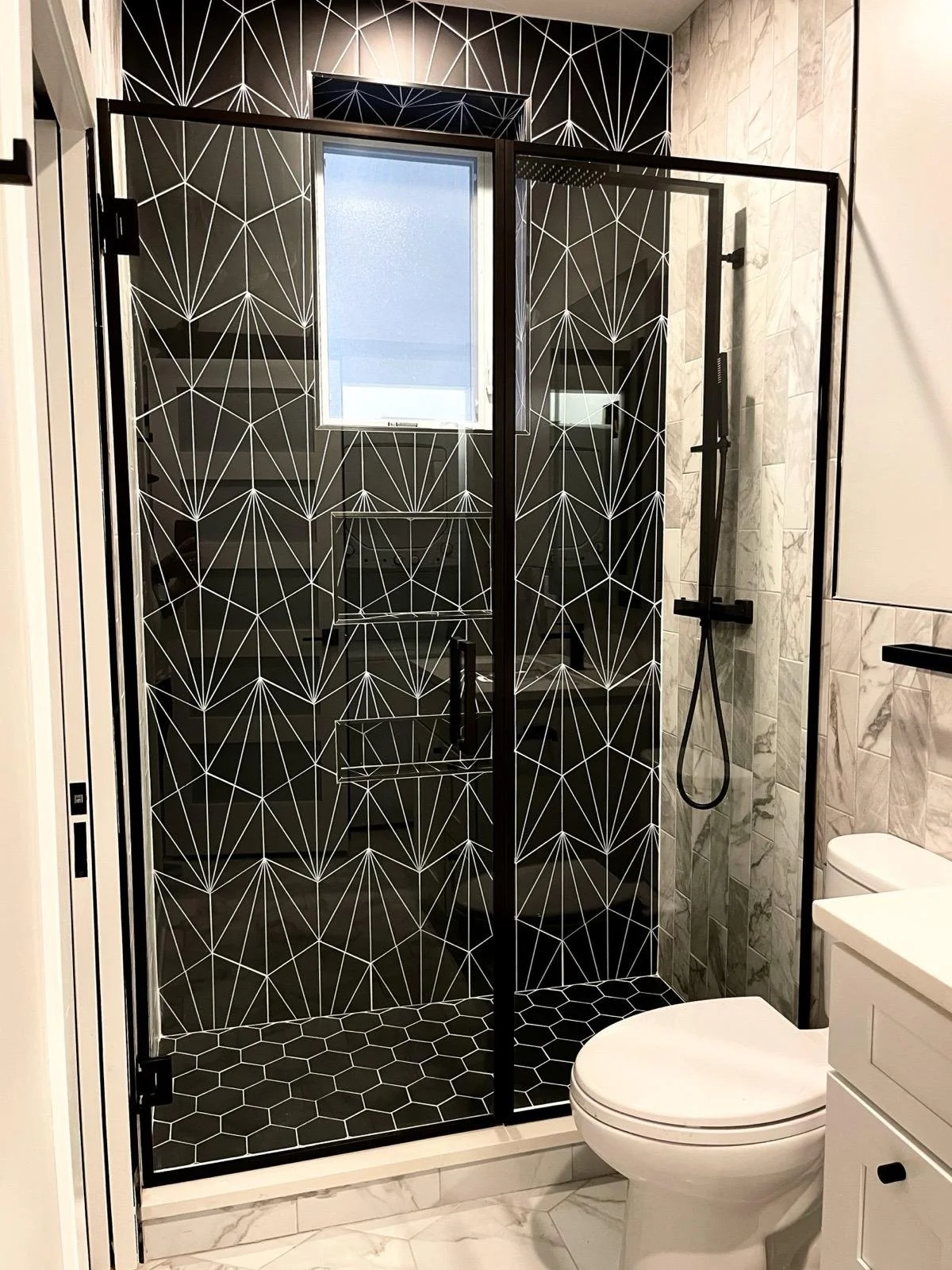 Modern bathroom with a glass shower enclosure featuring geometric black and white patterns, a window above the shower, a white toilet, a white vanity with black hardware, and marble flooring and wall tiles.