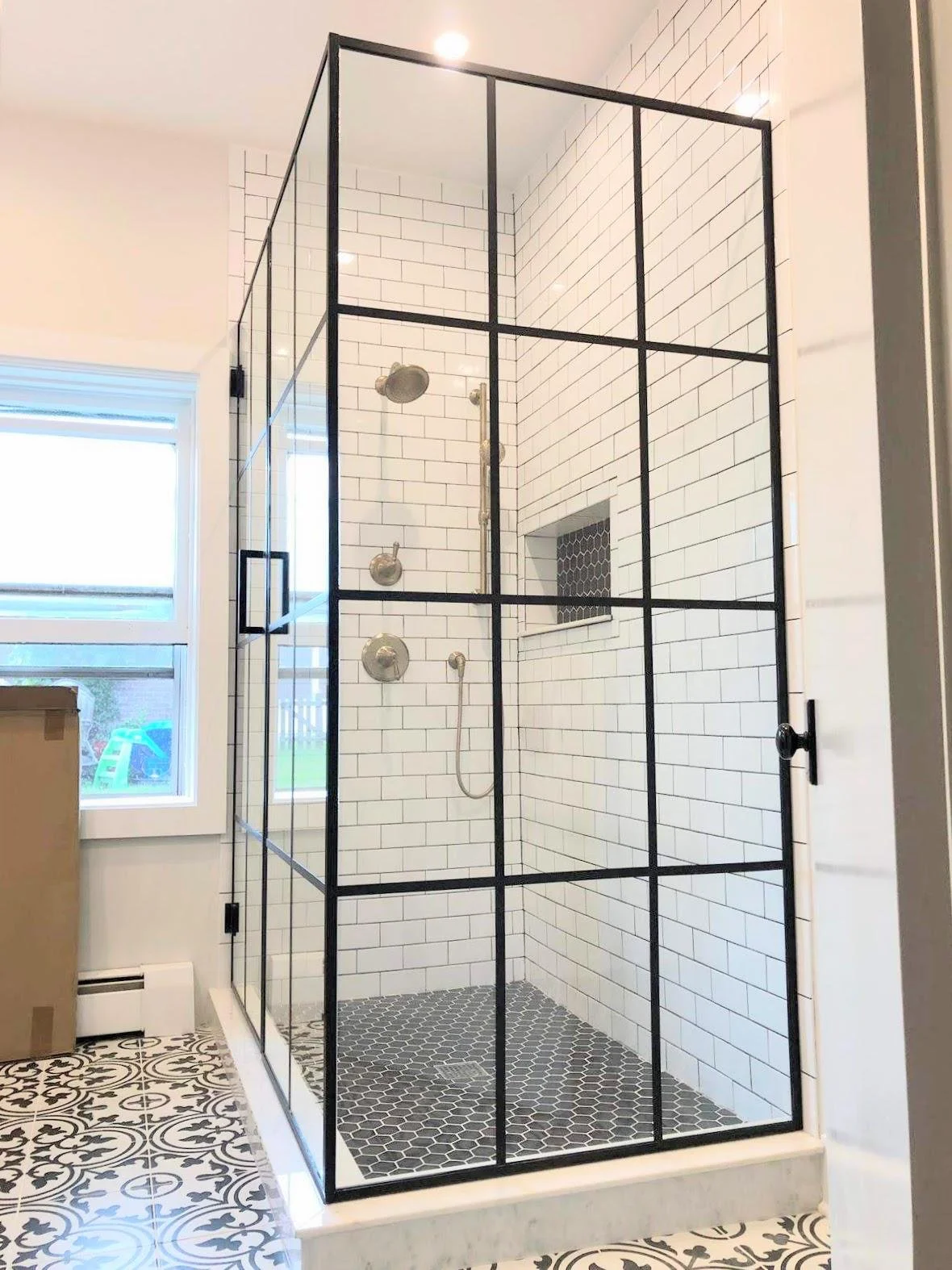 Modern walk-in shower with black metal frame, white subway tile walls, and black hexagonal tile flooring. Features a recessed shower niche and a rain showerhead, with a window nearby and patterned floor tiles outside the shower.