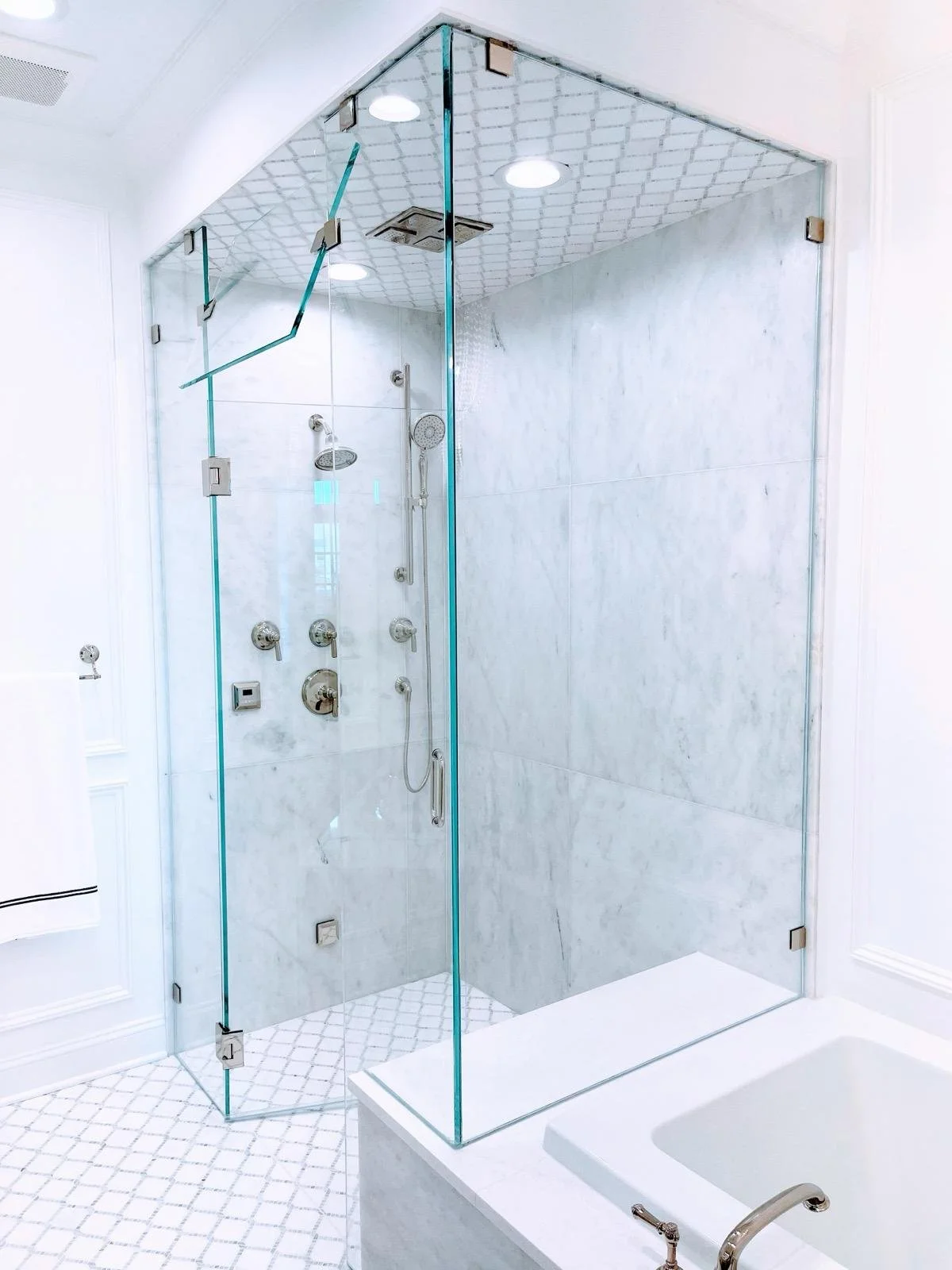 Modern glass-enclosed shower with multiple showerheads and controls, marble walls, and white tiled floor in a bathroom.