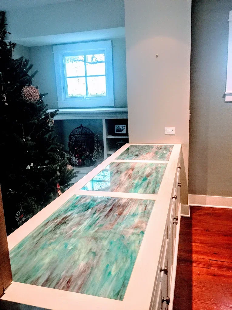 A kitchen counter with three glass top sections showcasing a colorful abstract design in shades of green, brown, and cream, with a Christmas tree and a window in the background.