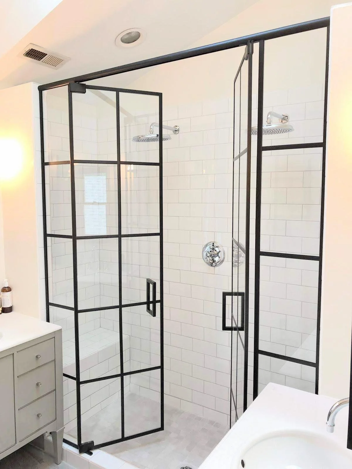 A modern shower with clear glass panels and black metal framing, featuring two rainfall showerheads on a white tiled wall, and a built-in bench inside the shower area.