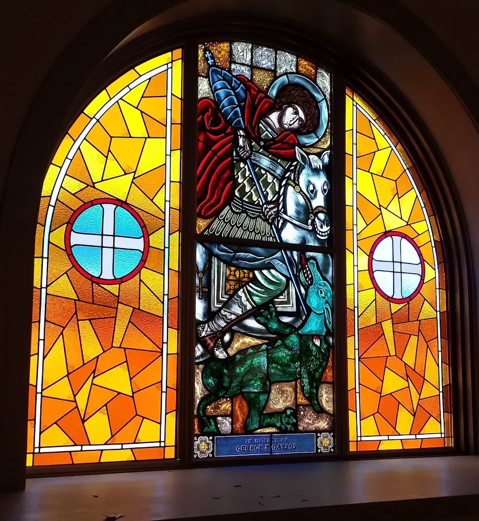 Colorful stained glass window featuring Saint George slaying a dragon, with yellow and orange background panels and circular blue and white accents.
