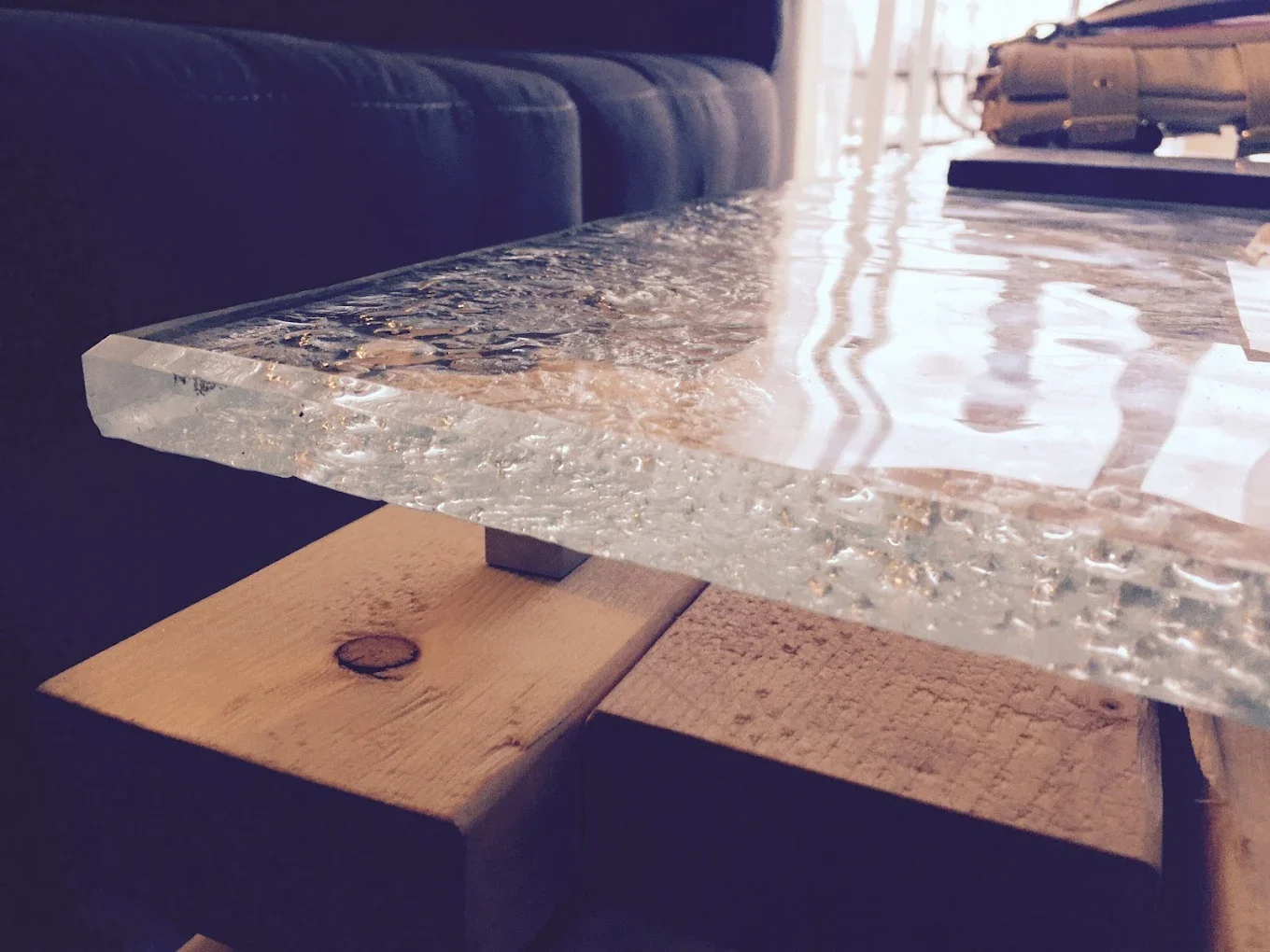 Close-up of a glass table with a textured surface, supported by wooden blocks, in a room with sunlight coming through windows, with a gray sofa in the background.