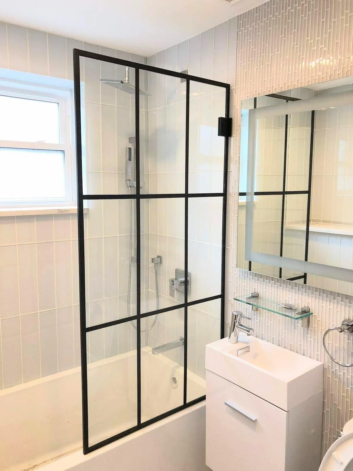 Bathroom with a walk-in shower enclosed by a black metal frame and glass, a small white vanity with a faucet, a mirror with a shelving unit, and a window