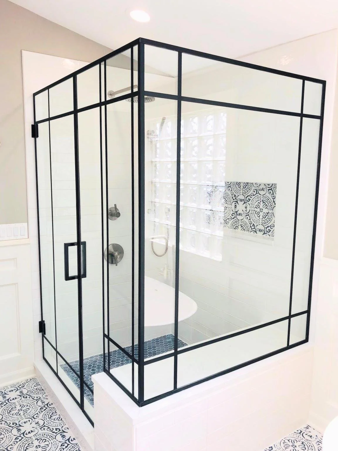 Frameless glass shower enclosure with black metal frame in a modern bathroom, featuring patterned and plain white tiles and a window with textured glass blocks.
