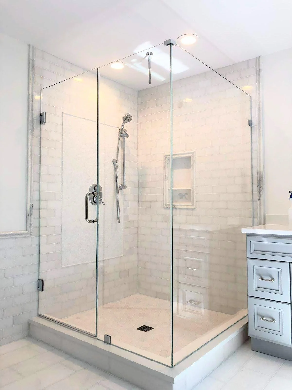 Bathroom shower with glass walls, chrome showerhead, and built-in shelf, next to white cabinetry.