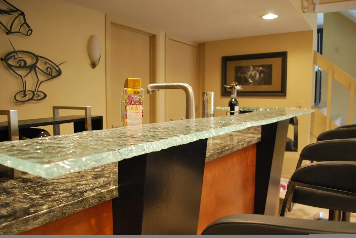 View of a kitchen counter with a glass countertop, a soap dispenser, a bottle of vodka, and a knife holder, with chairs and artwork on the walls in the background.