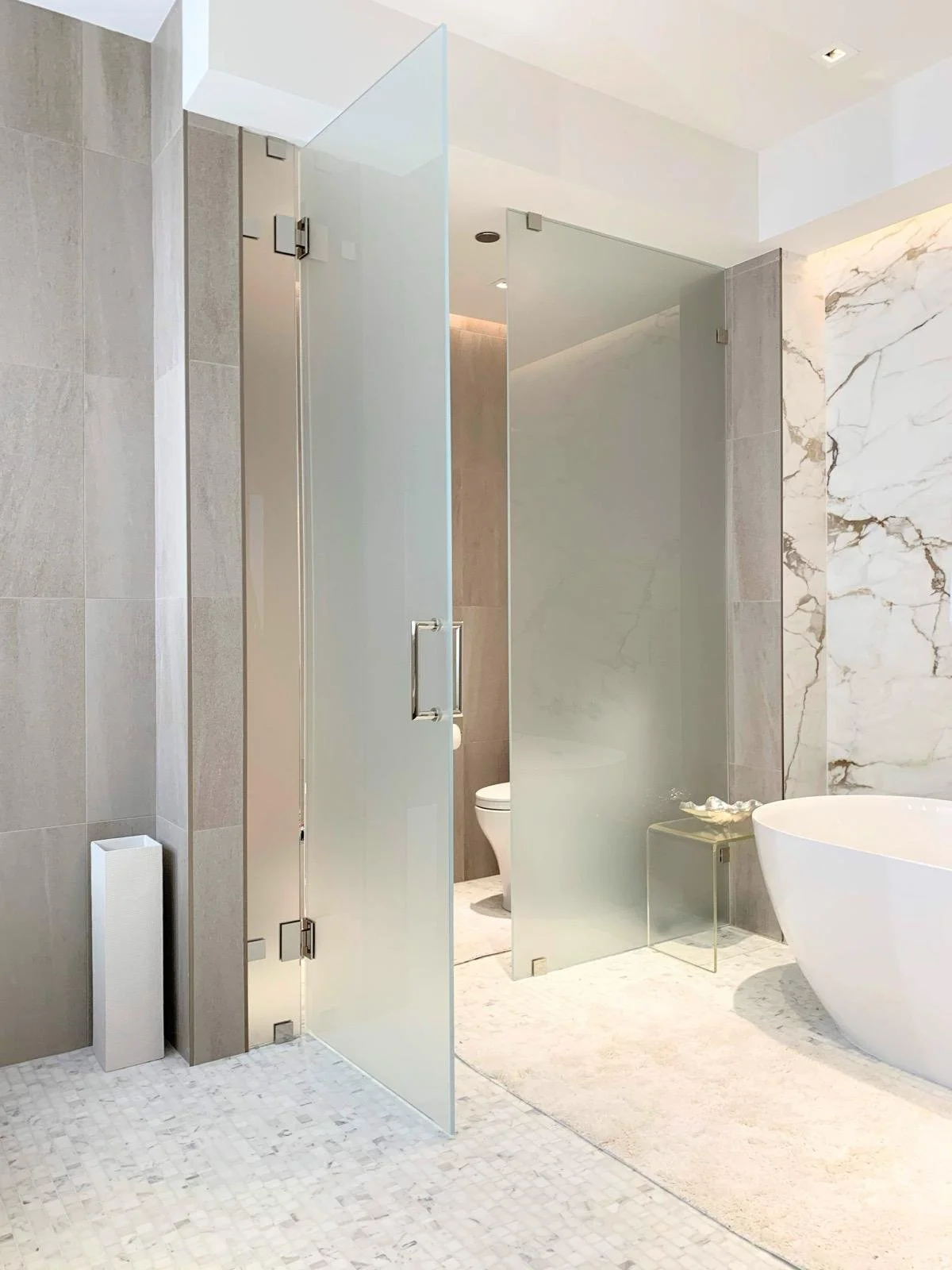 Modern bathroom with frosted glass walking shower, marble walls, and a white bathtub.