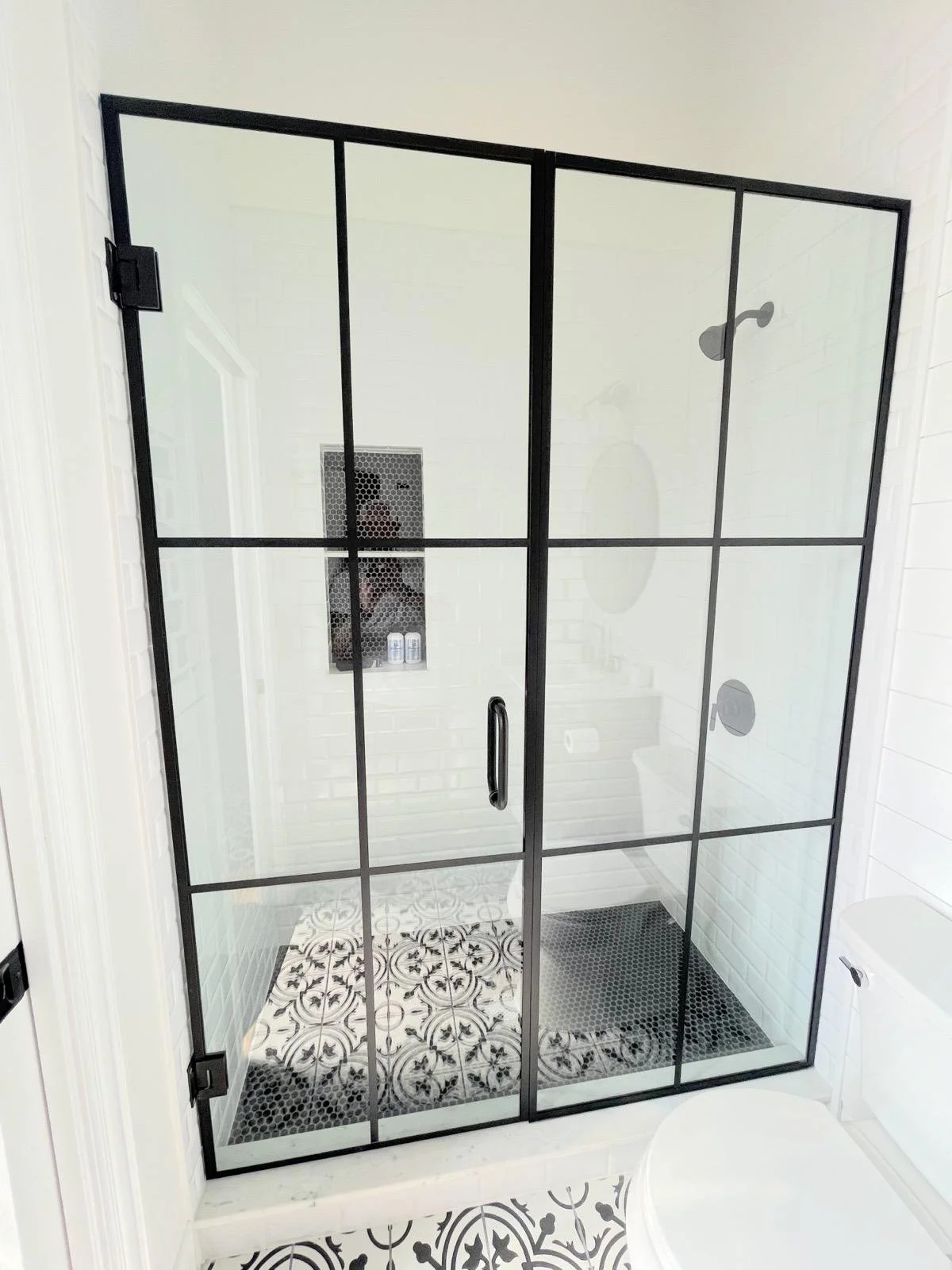 Modern shower with black-framed glass doors, black and white patterned floor tiles, white subway-tile walls, and a white bathtub nearby.