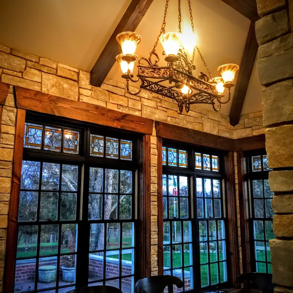 Interior view of a room with large windows featuring stained glass details at the top, stone walls, wooden framing, a chandelier hanging from the ceiling, and a view of an outdoor garden with trees.