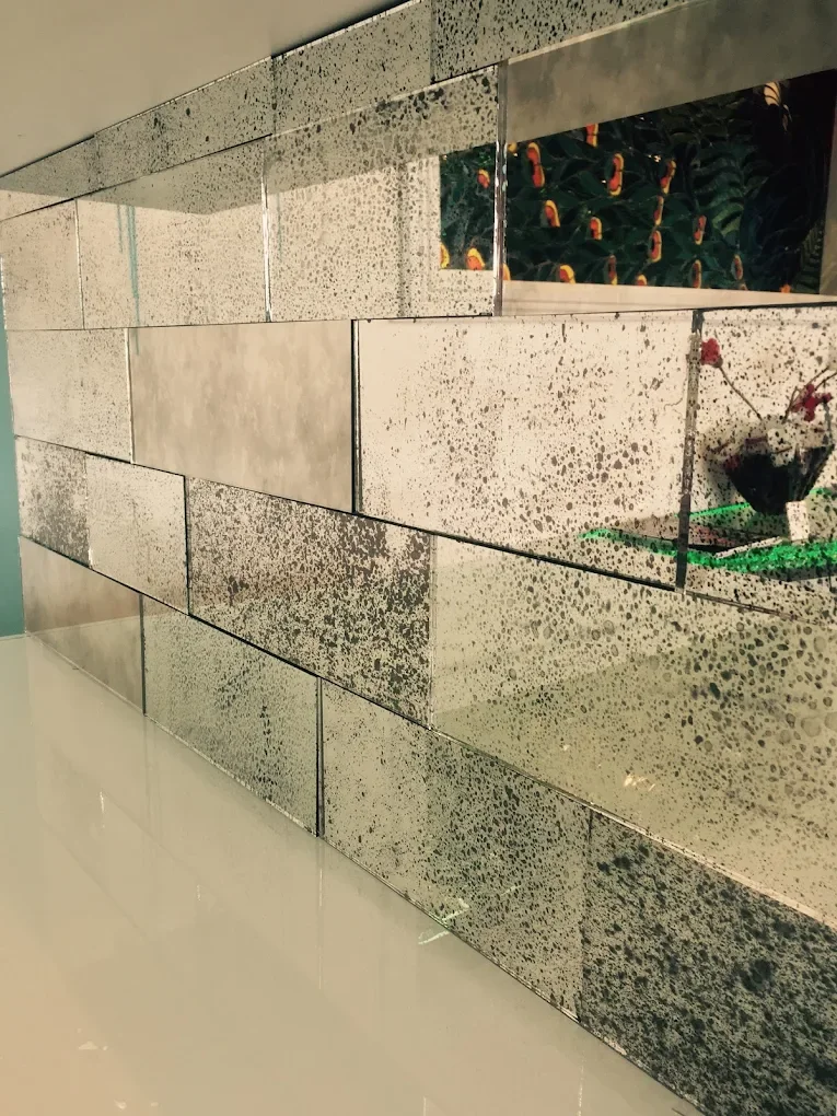 Close-up of a wall with metallic, speckled rectangular tiles arranged in a horizontal pattern, with a reflective surface showing a small vase with flowers and some decorative objects.