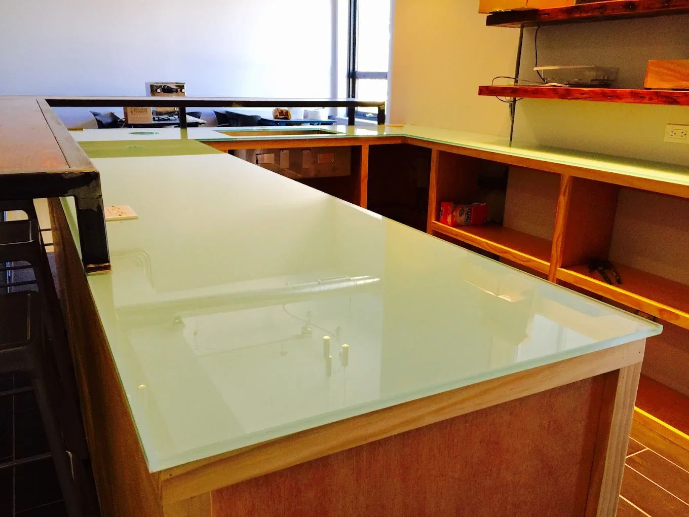 Empty retail or cafe counter with a glass top and wooden frame, with shelves behind it, in a well-lit room.