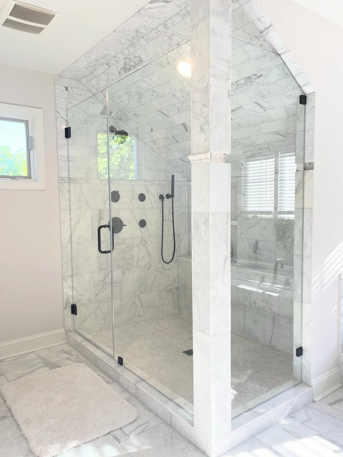 A modern walk-in shower with glass doors and marble tile walls and ceiling, featuring black fixtures, a hand-held showerhead, and a built-in bench; a window with blinds and a plush white rug outside the shower.