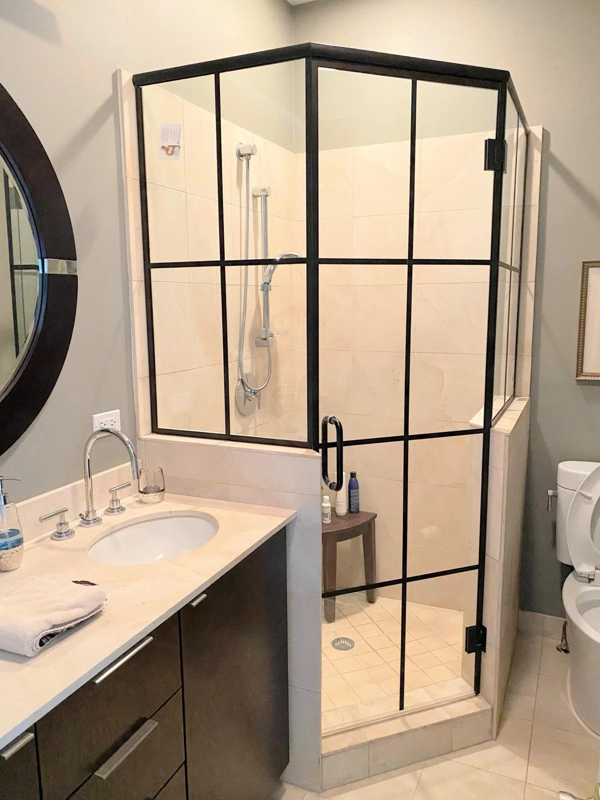 A bathroom with a shower enclosure with black metal framing, a glass door, beige tiled walls, a dark vanity with a white countertop, a round mirror, and a toilet to the right.