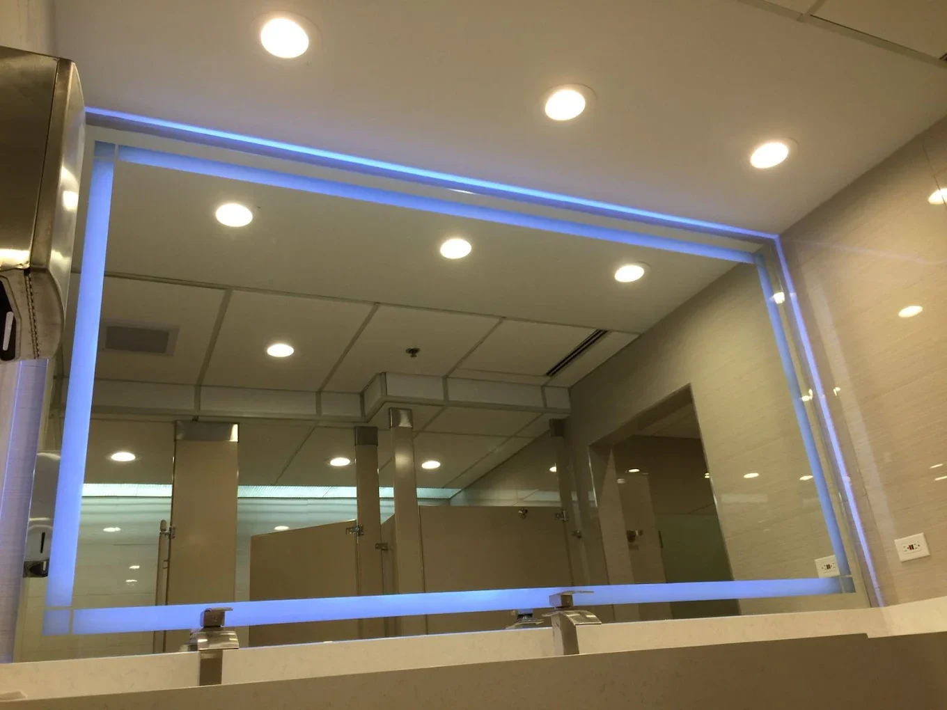 A large mirror with blue LED lighting border above a white countertop in a bathroom. The mirror reflects ceiling lights, partitions, and bathroom stalls.