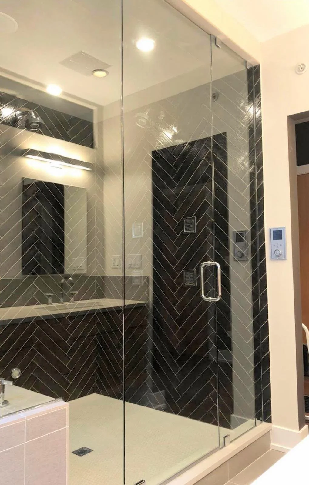 A modern glass shower enclosure with black tiling in a bathroom. Inside the shower, there are two black wall-mounted showerheads and a stainless steel handle on the glass door. The bathroom features a wall-mounted mirror, a white sink with a faucet, 