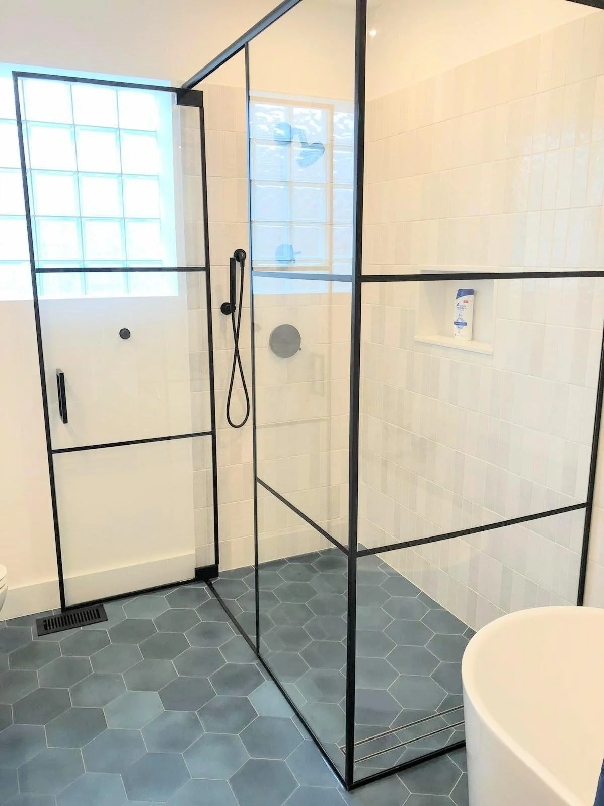 Modern walk-in shower with glass walls and black metal frame, white tiled walls, grey hexagon floor tiles, and a built-in corner shelf holding a bottle of body wash.