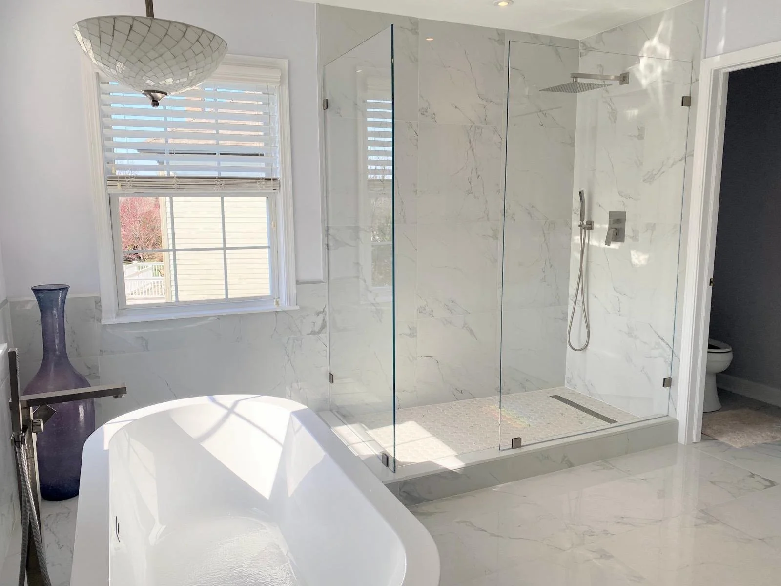 Modern bathroom with a white bathtub, a window with blinds, a glass-enclosed walk-in shower with marble walls, and a toilet in a separate room.