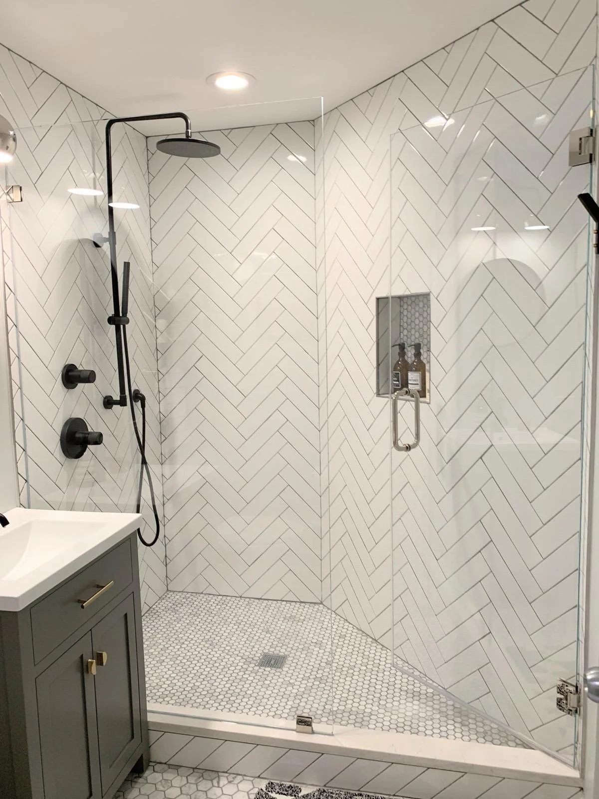 Modern bathroom with walk-in shower featuring white herringbone tiles, a built-in shampoo niche, black shower hardware, and a gray vanity with a white countertop.