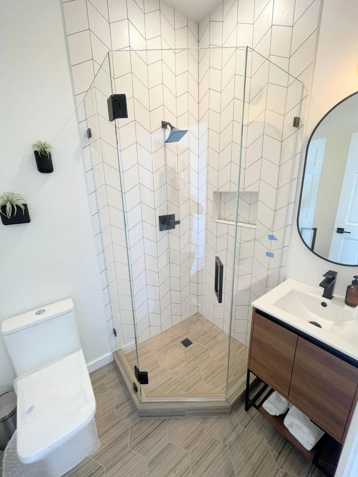 Bathroom with a glass shower enclosure, white hexagonal wall tiles, a white toilet, a wooden vanity with a white sink, a round mirror, and small wall-mounted plant pots.