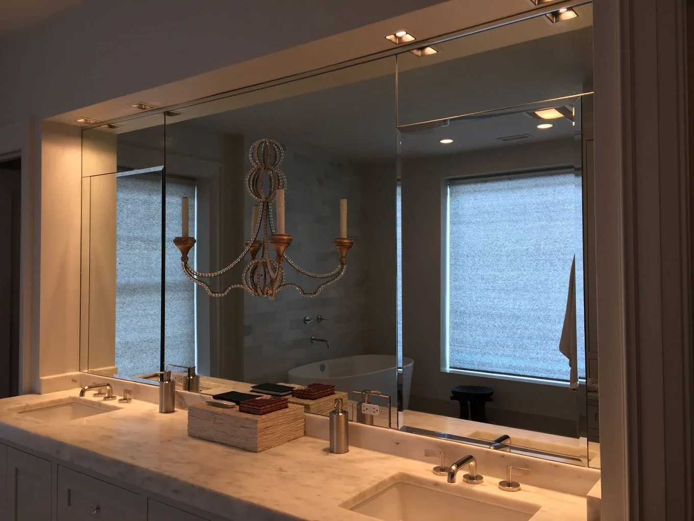 A bathroom with a marble countertop, double sinks, and a large mirror. There are two soap dispensers, a tray, and some items on the counter. A chandelier hangs from the ceiling. In the background, there is a bathtub with a wall-mounted faucet, a wind