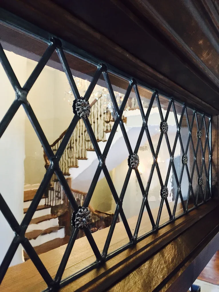 View of a decorative black metal grille with floral accents, reflecting a staircase and interior room.
