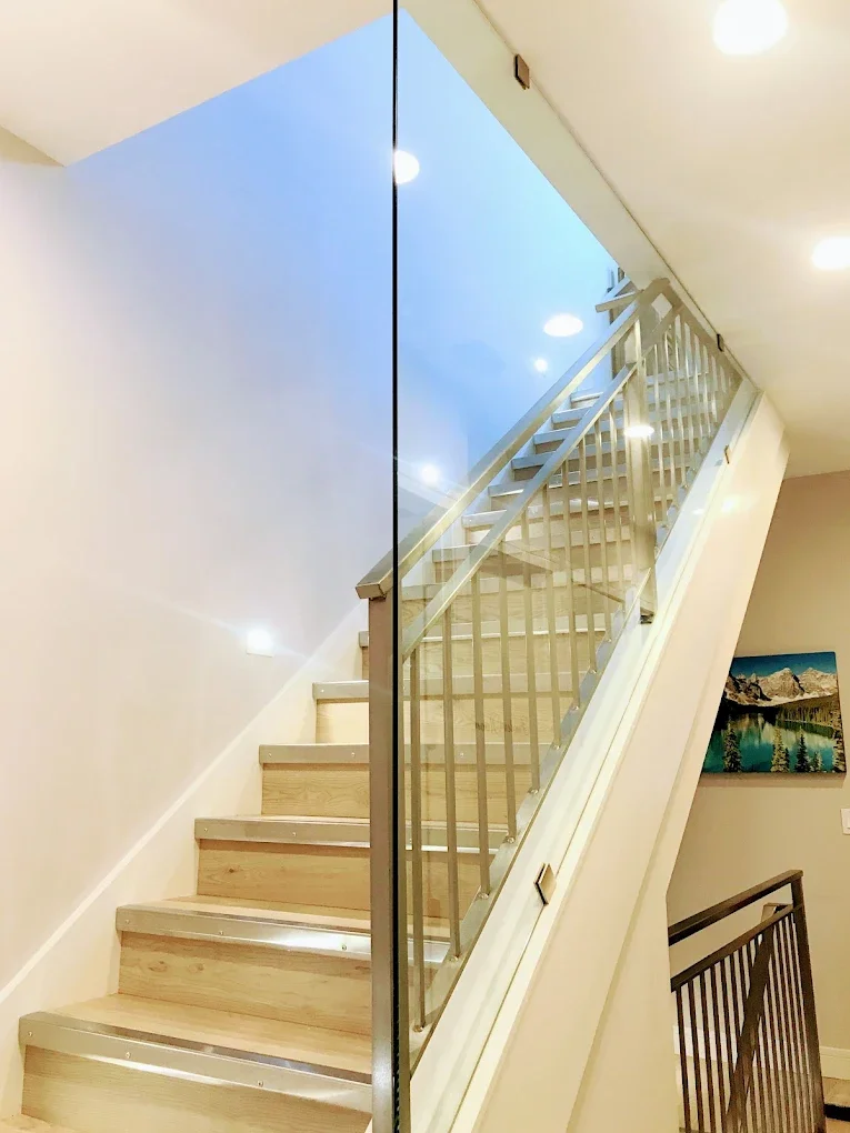 Interior staircase with wooden steps and glass railing leading upwards, with a painting of mountains and water on the wall nearby.