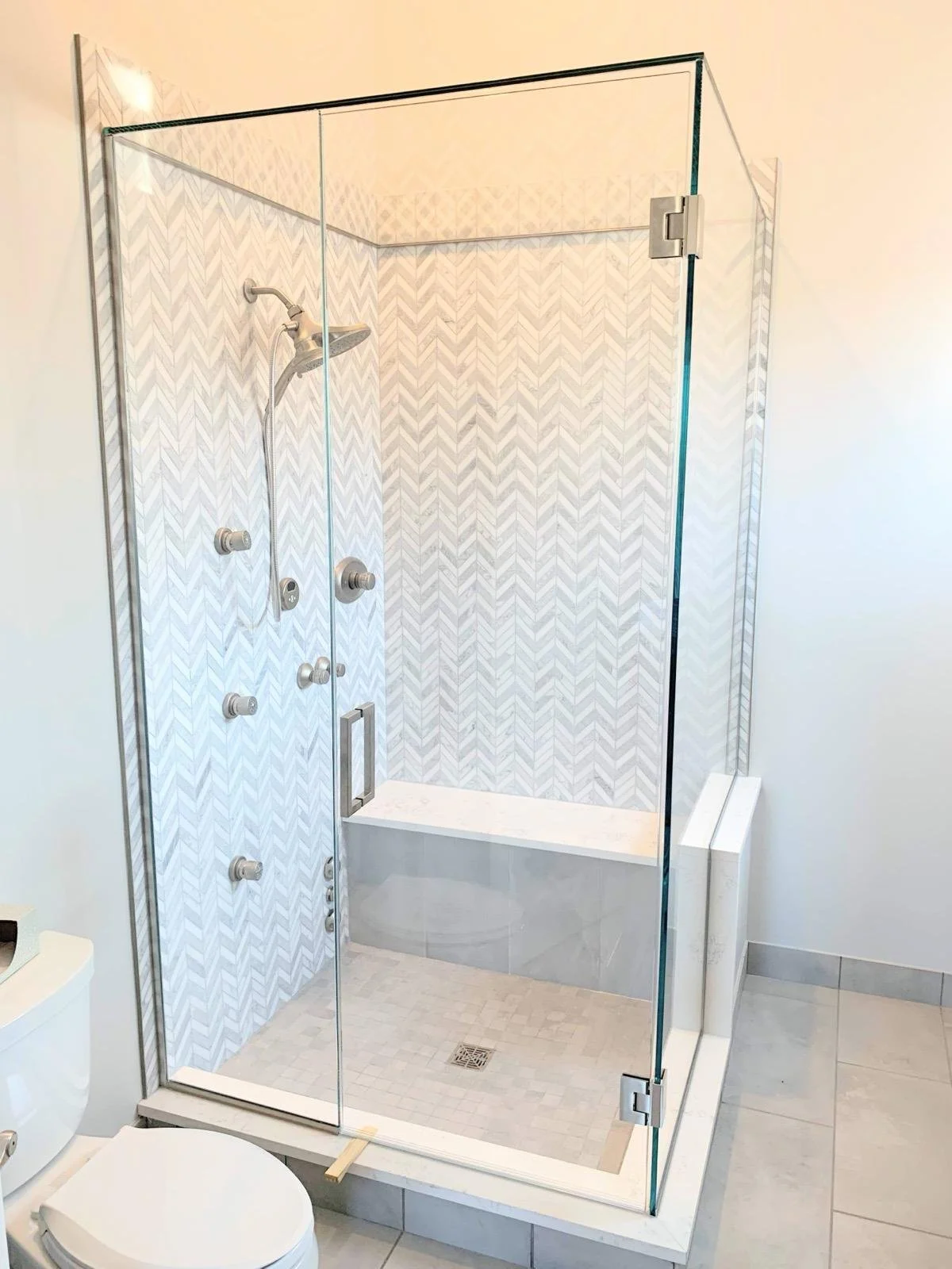 A modern glass-enclosed shower with a zigzag patterned wall inside, a built-in bench, and multiple shower controls. Part of a bathroom with tiled floor and a white toilet visible in the foreground.
