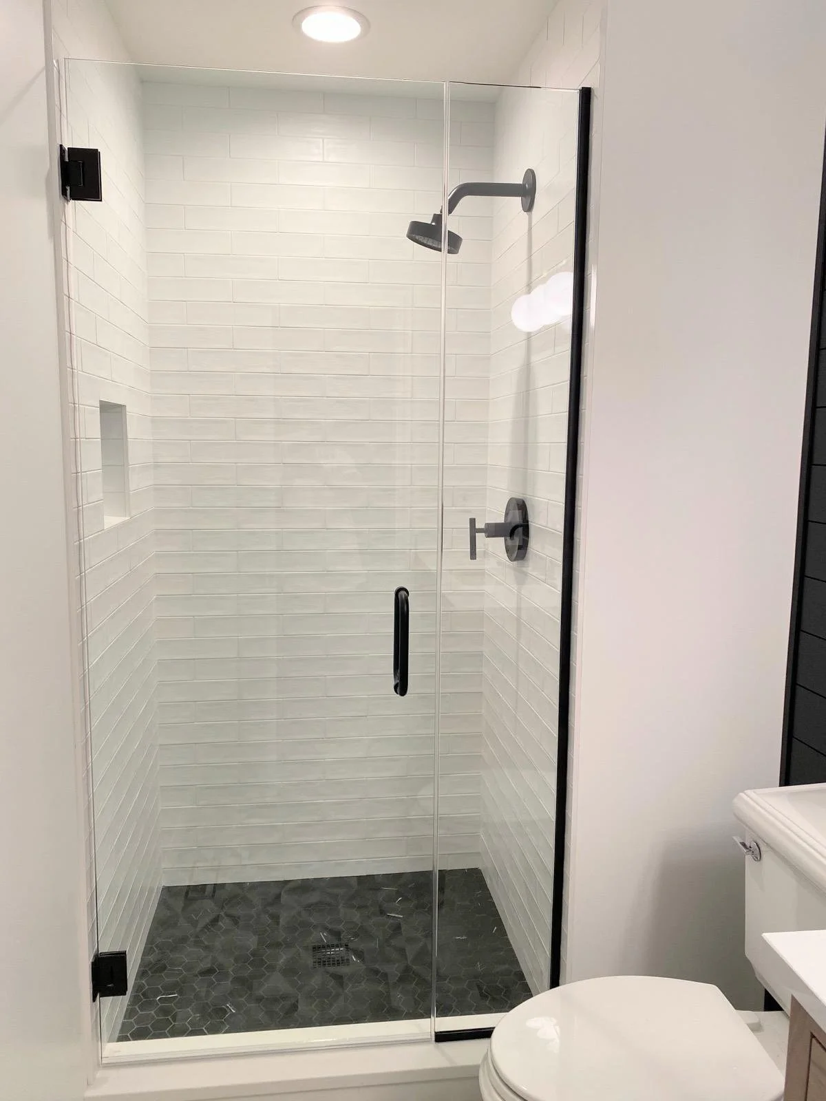 A modern walk-in shower with a glass door, white subway tile walls, a black shower head and handle, and black hexagonal floor tiles, alongside a white toilet.