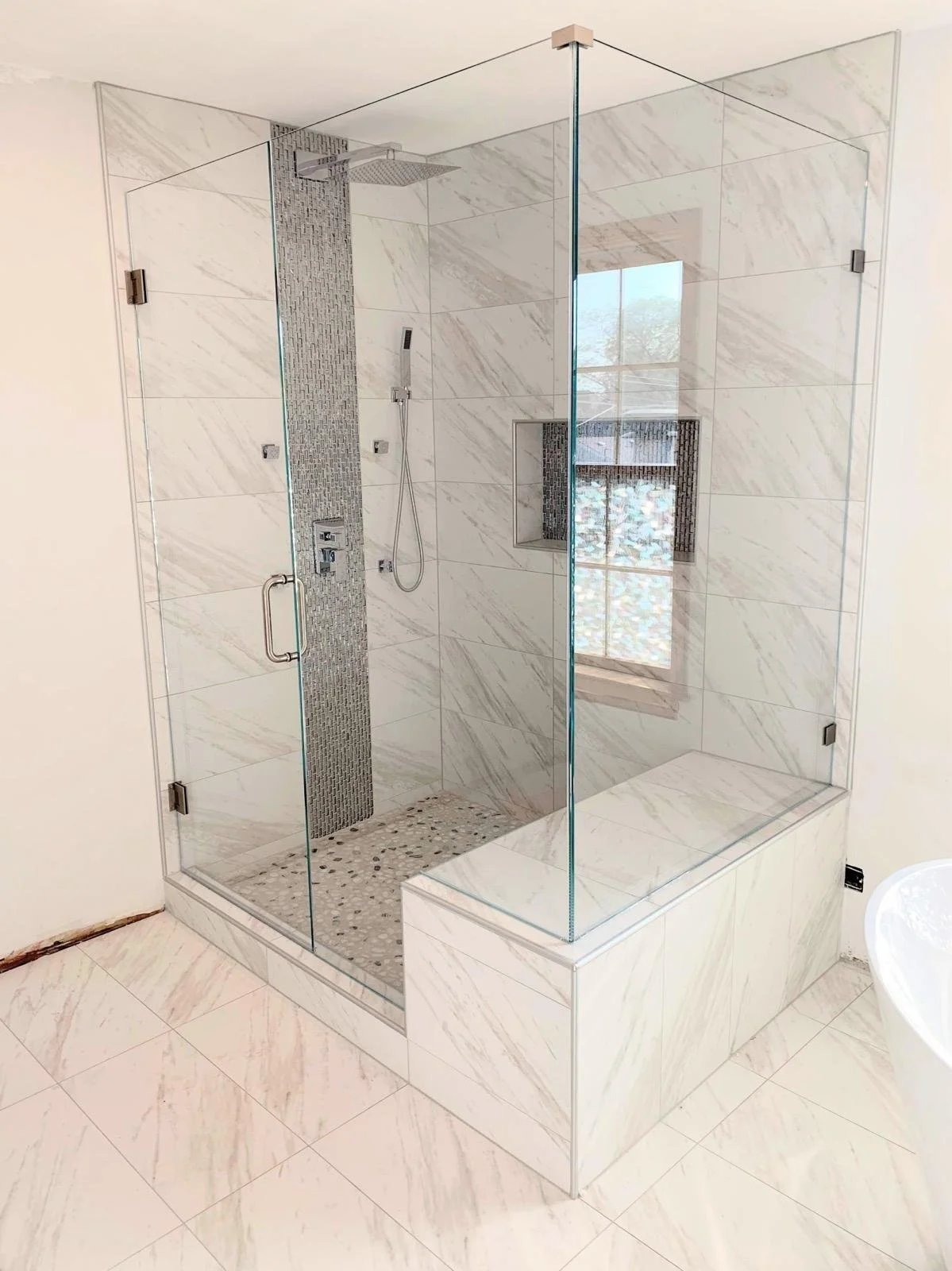A modern shower area with glass walls, marble tiles, a rainfall showerhead, and a built-in niche with black mosaic tiles, adjacent to a window.