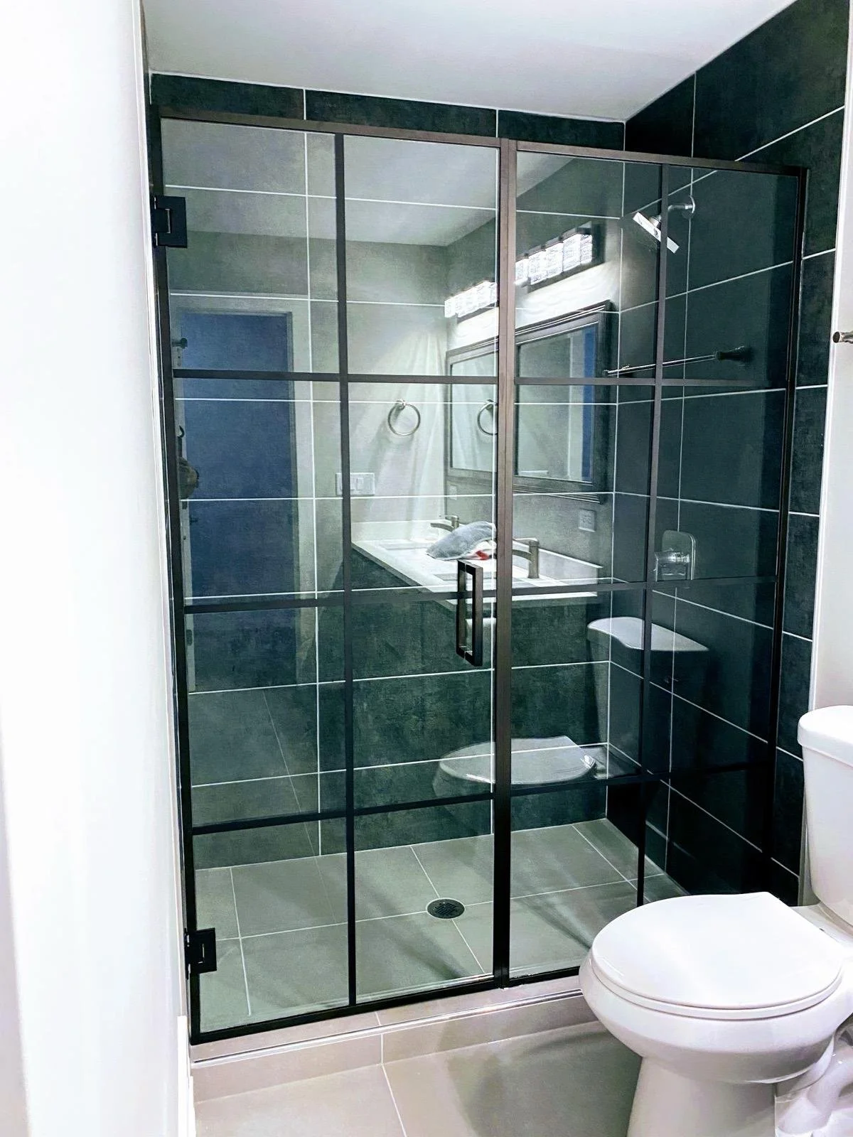 Modern bathroom with a glass shower enclosure, dark green tile walls, a white toilet, and a vanity with a mirror and lighting above.