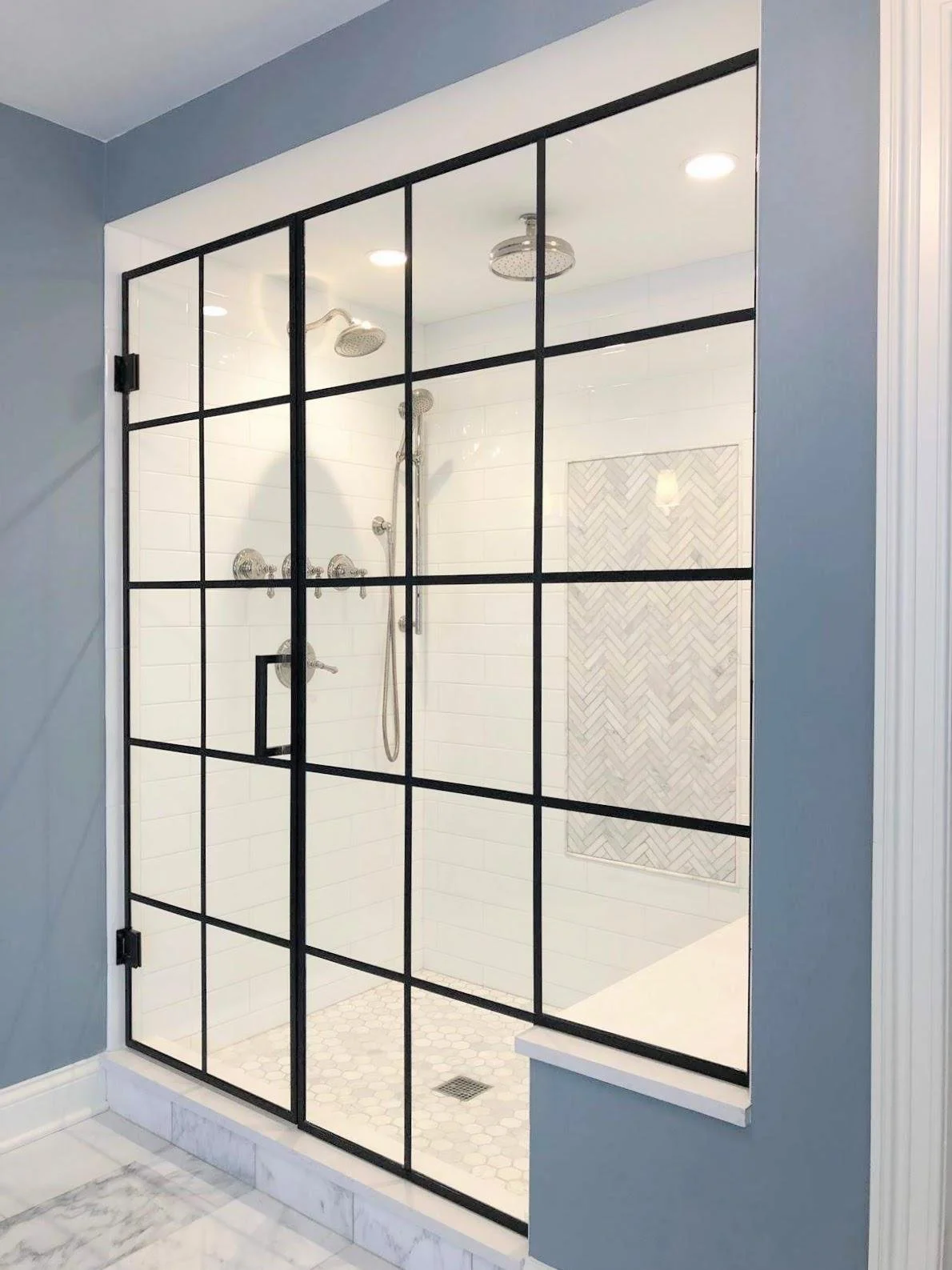 View of a modern walk-in shower with black-framed glass doors and white subway tile walls, featuring double showerheads and a built-in niche with herringbone tile backsplash.