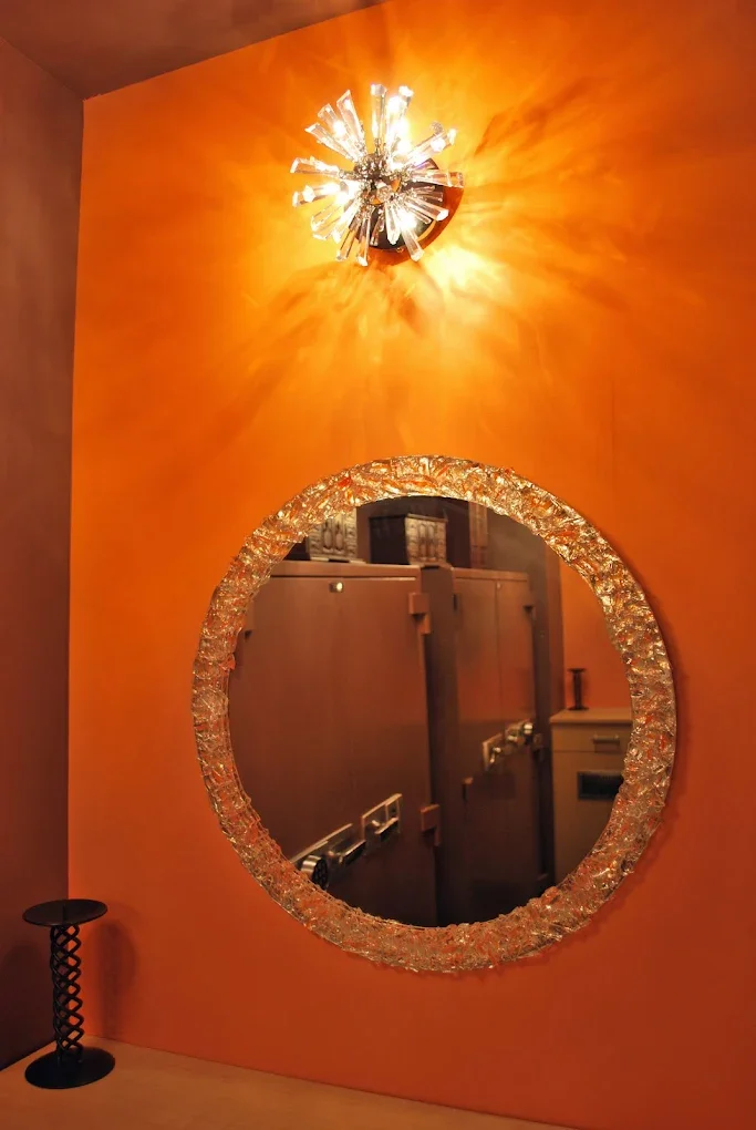 Decorative mirror with a textured, glittering frame hanging on an orange wall, with a modern, shiny ceiling light above it emitting bright light and shadows.