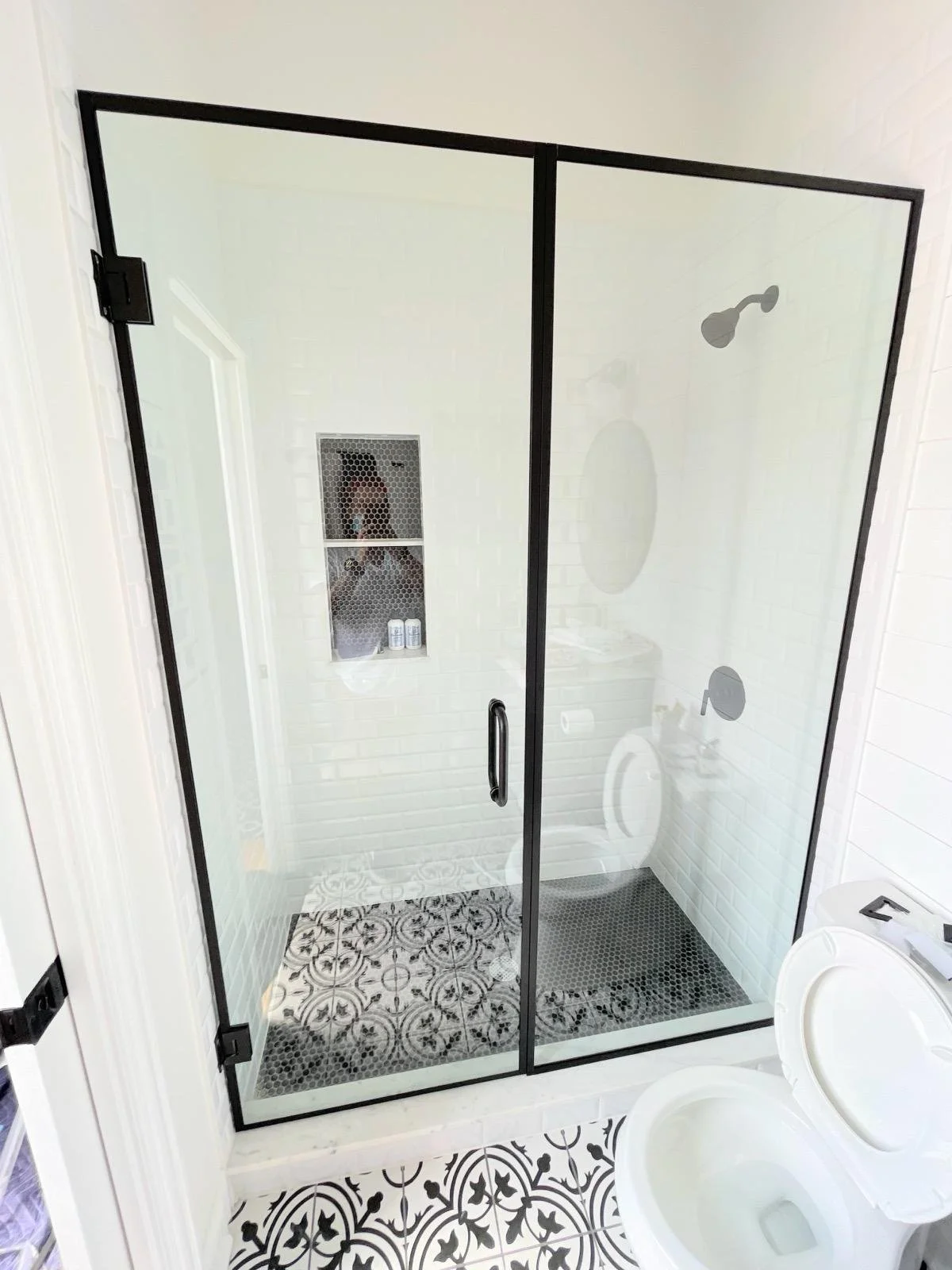 Bathroom with a glass shower enclosure, patterned black and white floor tiles, white walls, and a toilet with an open lid.