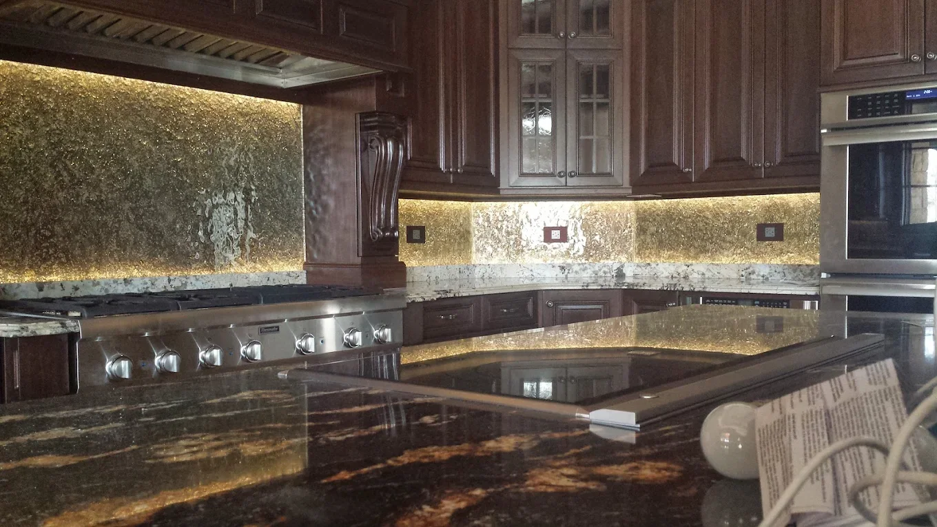 A kitchen with dark wood cabinets, gold and brown marble countertops, and a matching gold and brown marble backsplash. Visible appliances include a stainless steel stove and oven, with electrical outlets on the wall.