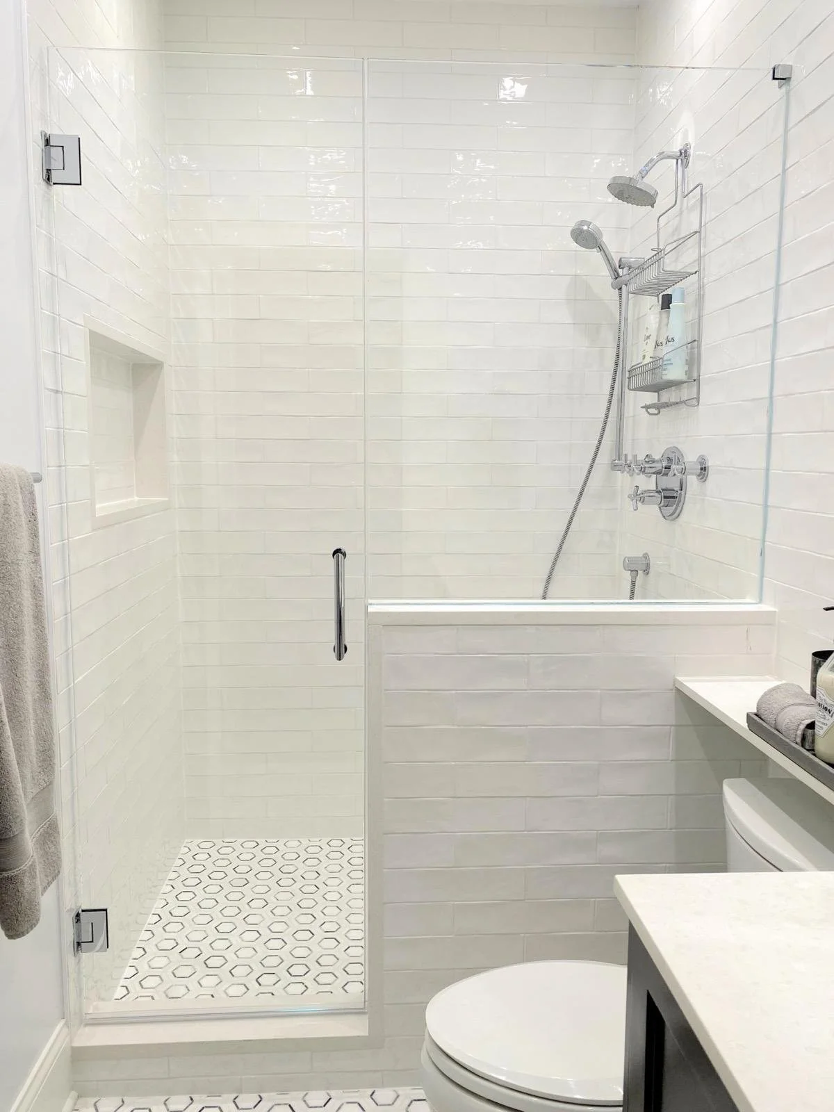 Modern white bathroom with a glass-enclosed shower featuring white tiled walls and patterned floor tiles, and a white toilet.