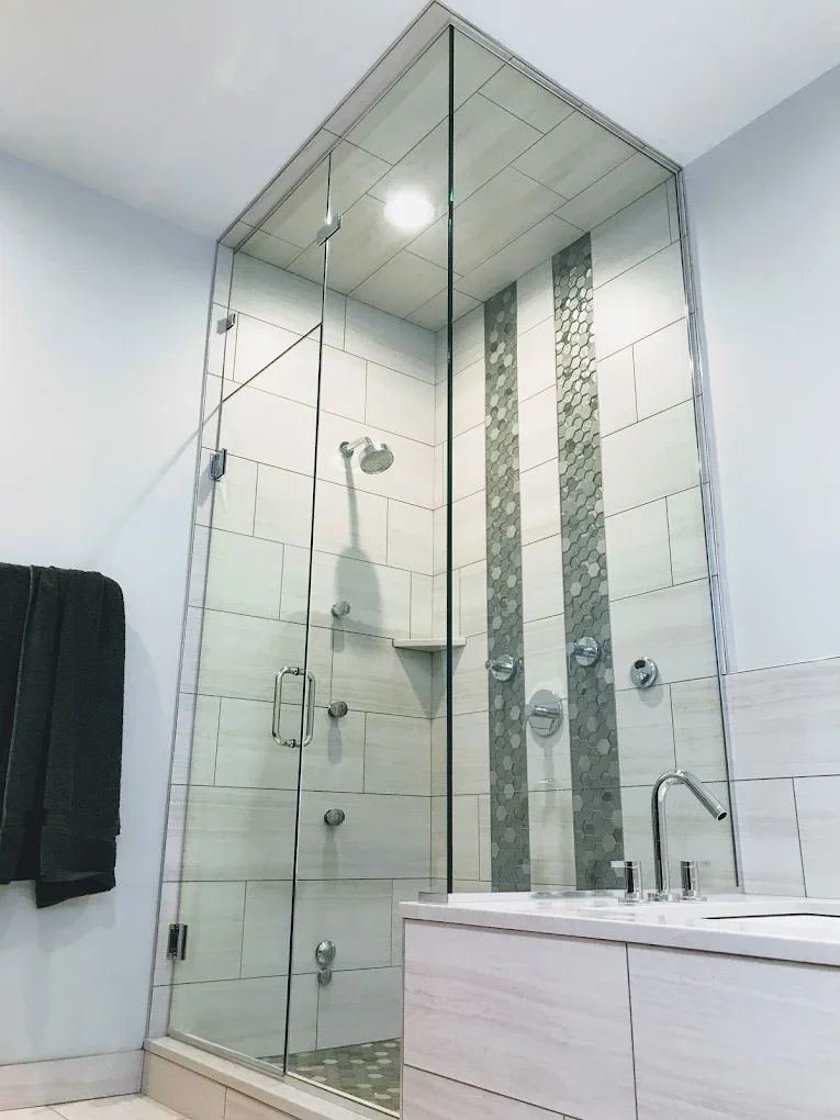 Modern bathroom with a glass shower enclosure and a bathtub beneath it, decorated with vertical gray mosaic tiles, a black towel hanging on a white wall.
