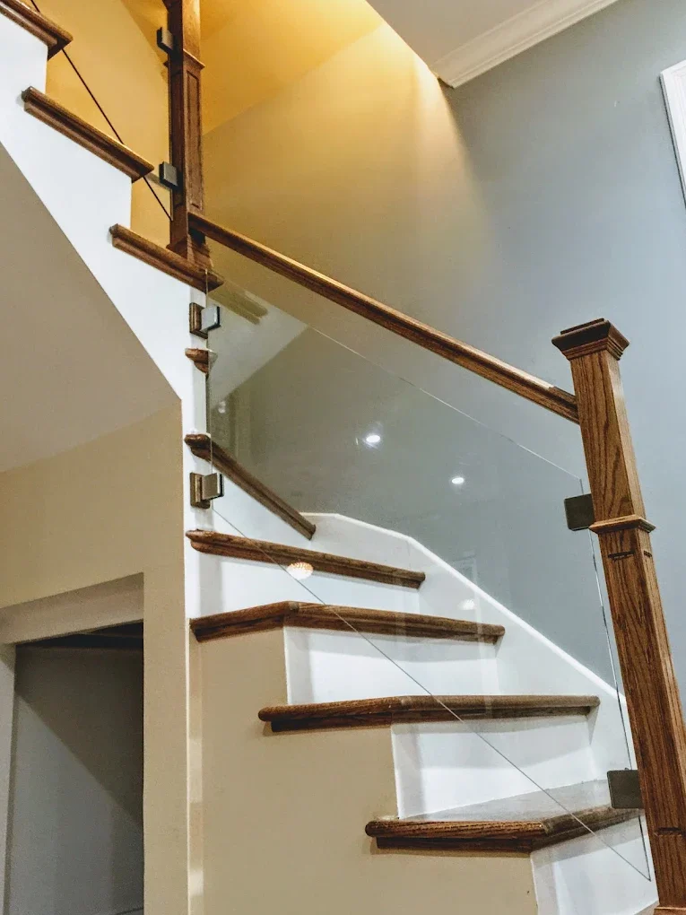 Interior view of a staircase with wooden steps and a glass railing, inside a home with beige walls and ceiling lighting.