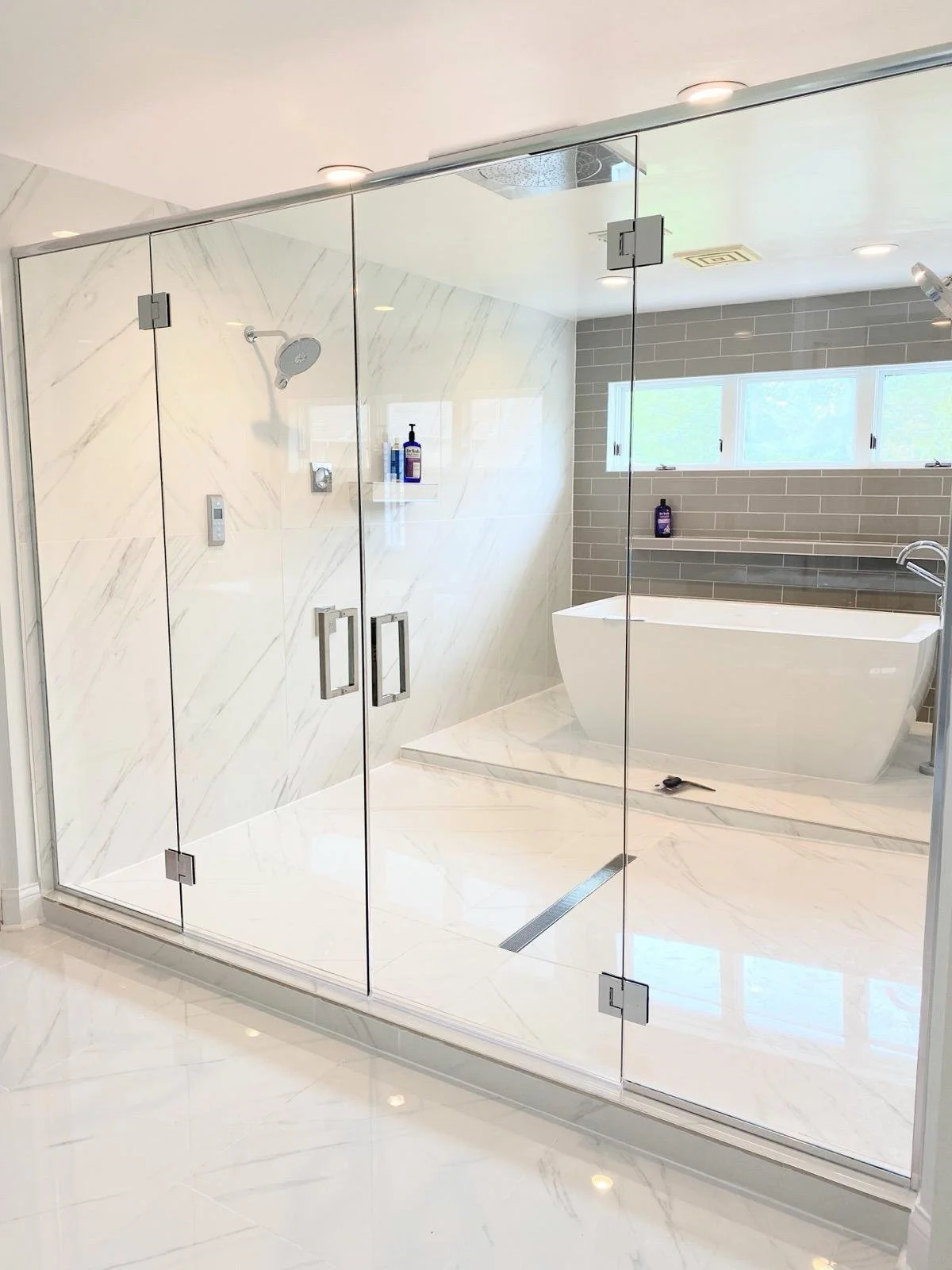 Modern bathroom with a glass-enclosed shower area and a bathtub, done in white marble and gray tiles, with windows letting in natural light.