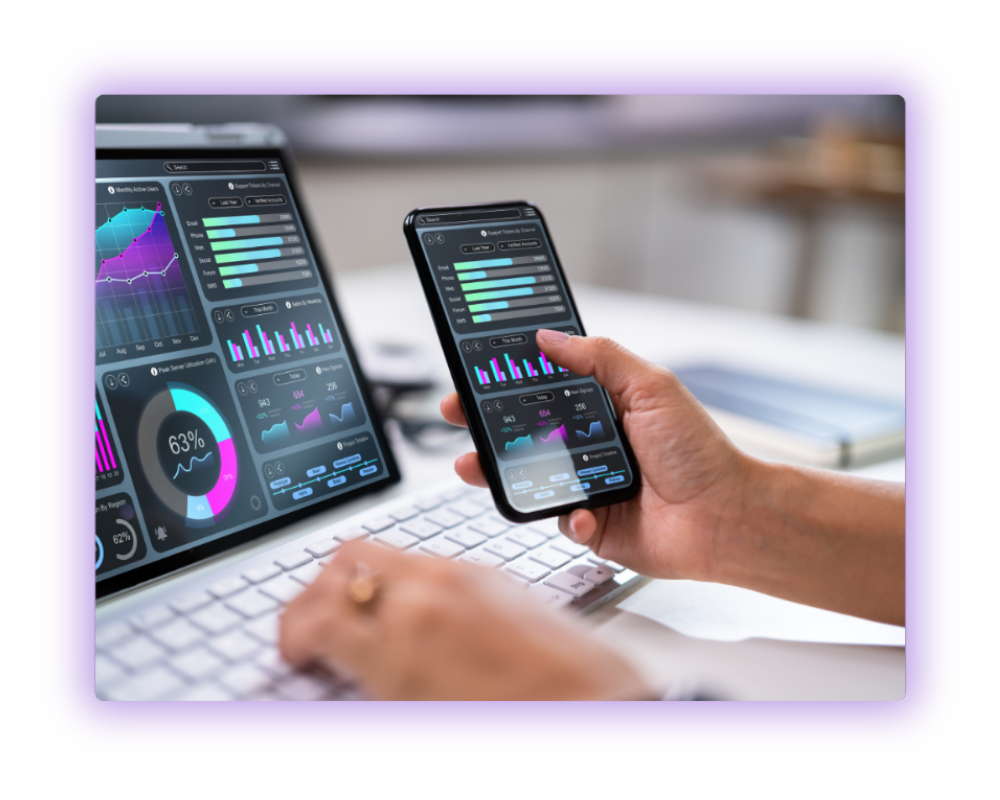 A person using a smartphone and laptop to view data analytics dashboards with graphs and charts on both screens.