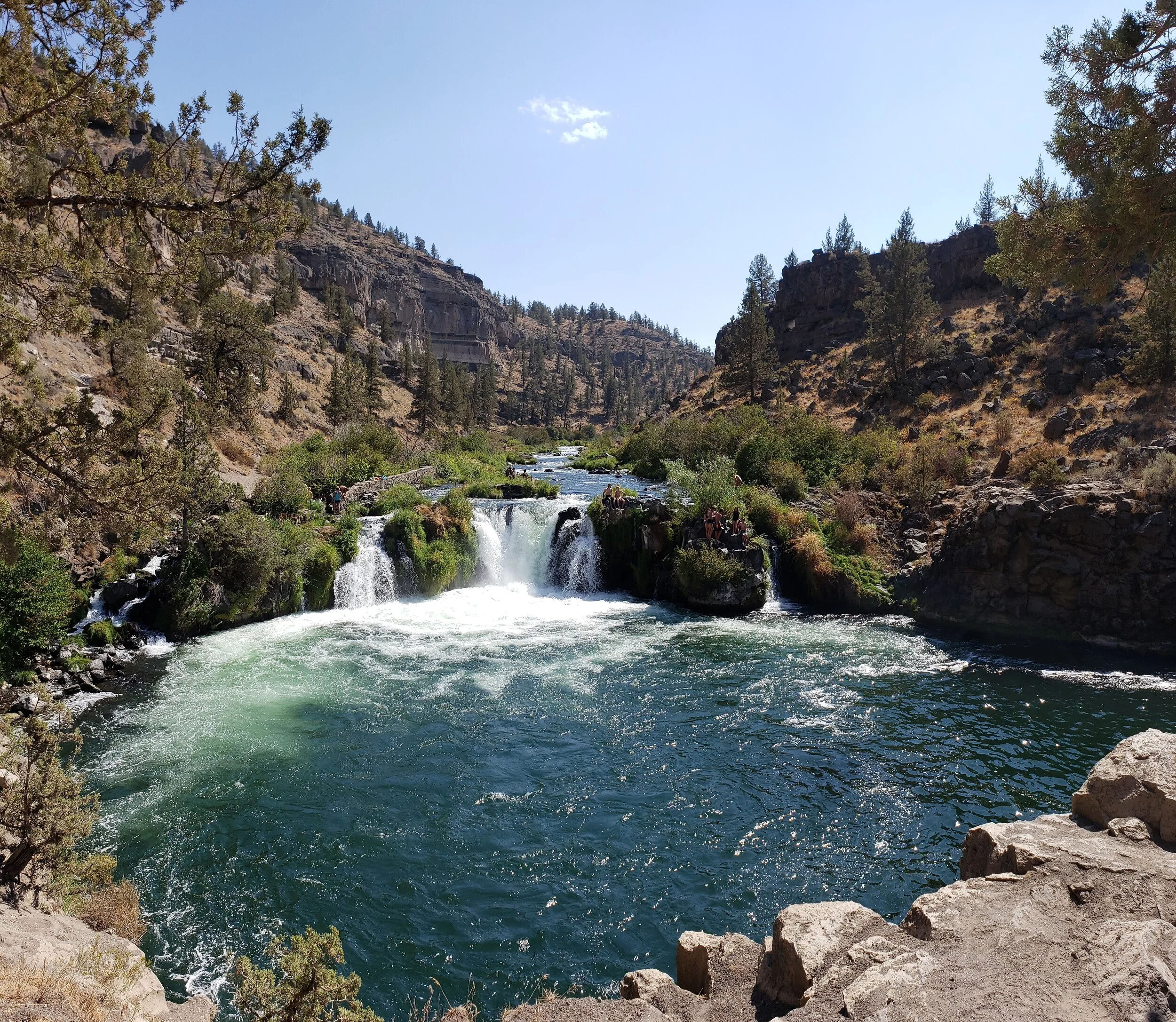 A scenic river with small waterfalls flowing through a canyon, surrounded by rocky cliffs and sparse trees under a clear blue sky.