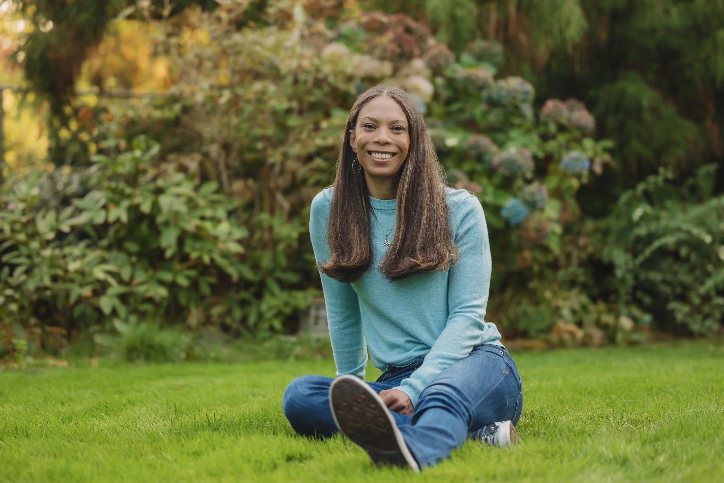 A woman sitting on a grassy lawn smiling at the camera, with a background of trees and bushes.