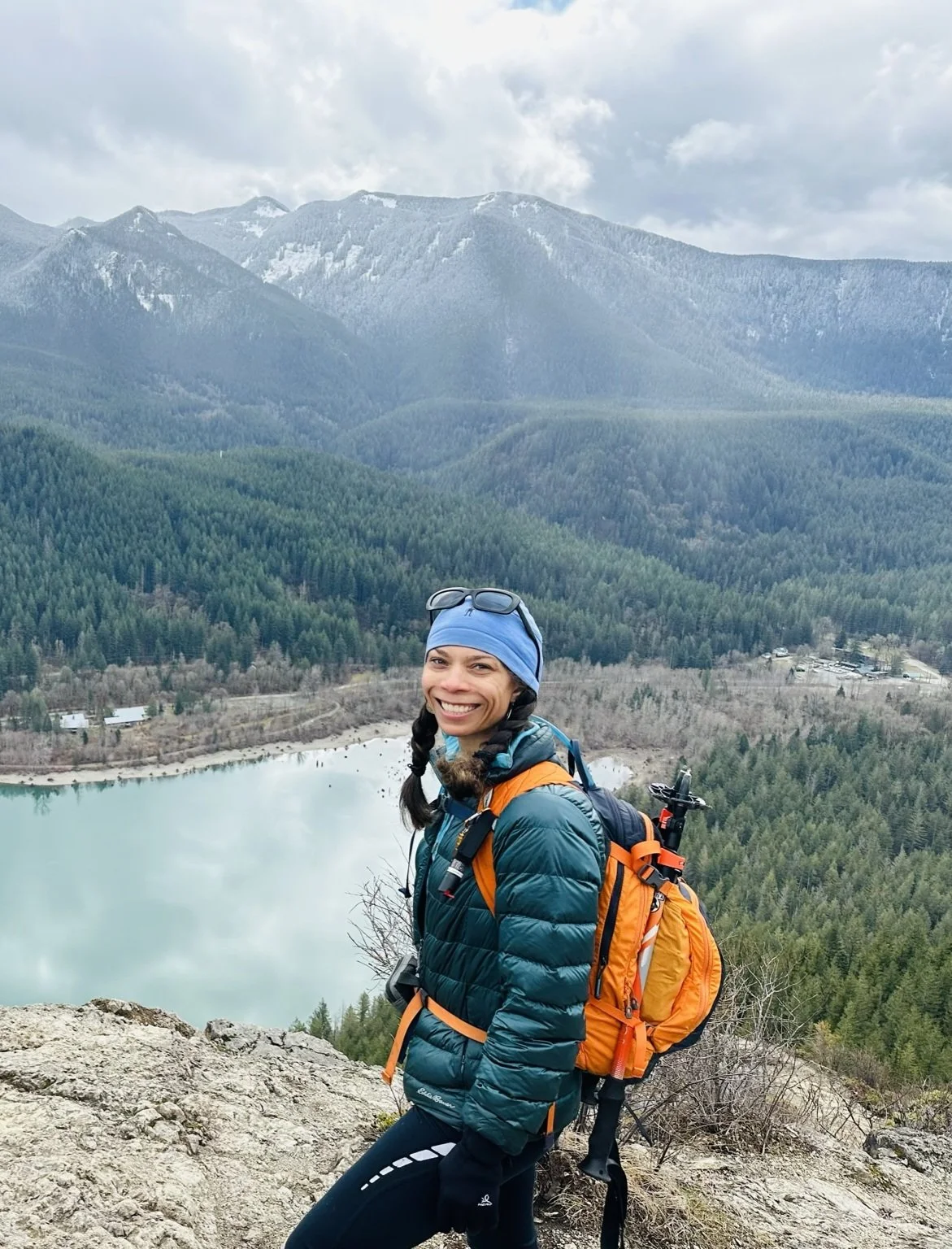 Woman in outdoor gear smiling on a mountain overlooking a lake and forested hills with mountains in the background.