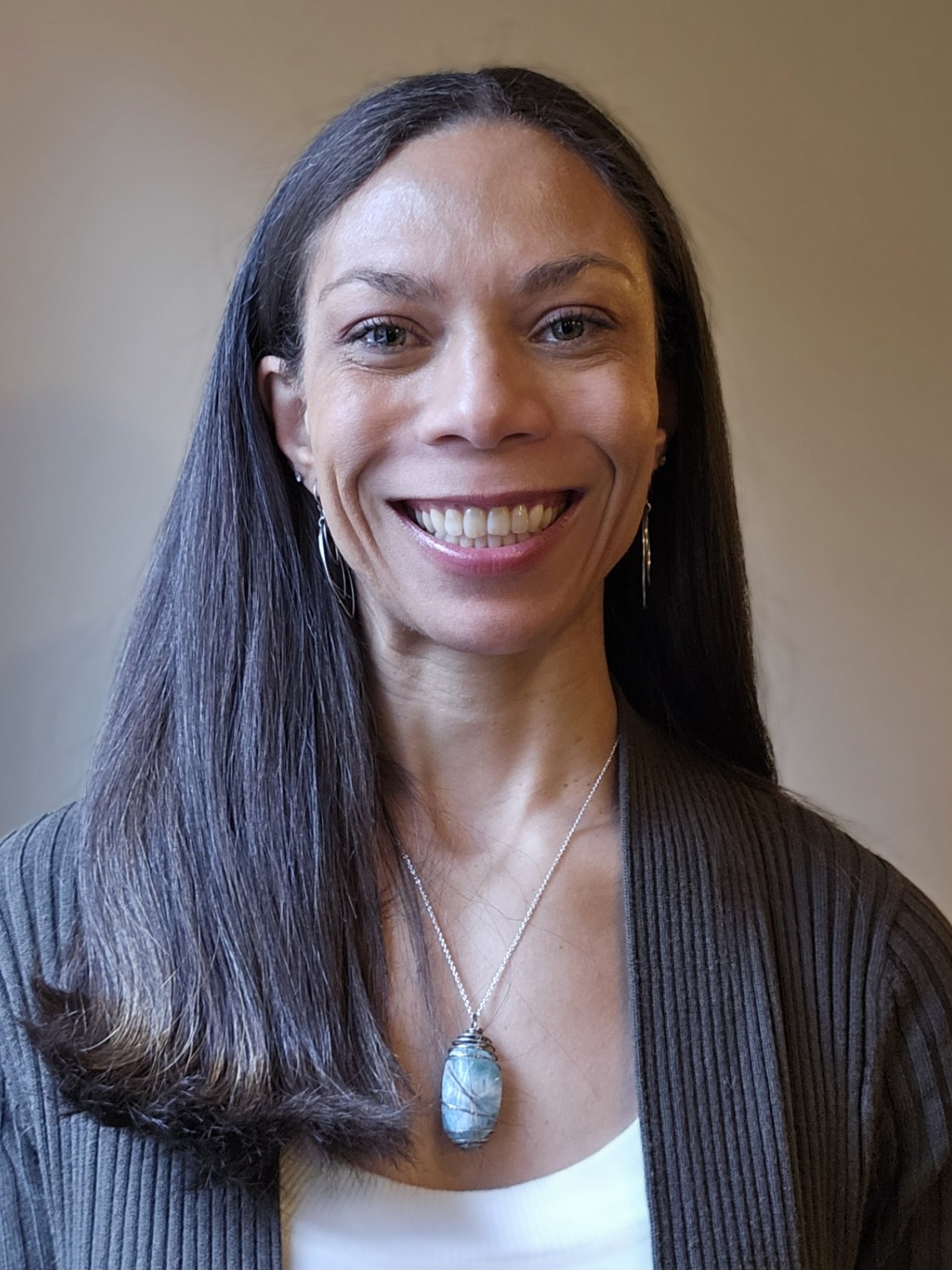 A woman with long dark hair wearing a necklace with a large pendant, earrings, a white top, and a brown blazer, smiling at the camera.