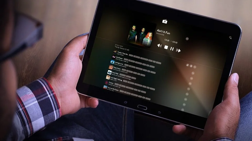 Person holding a tablet playing a music video or song titled 'Ain't Fun' from the playlist with various song titles visible.