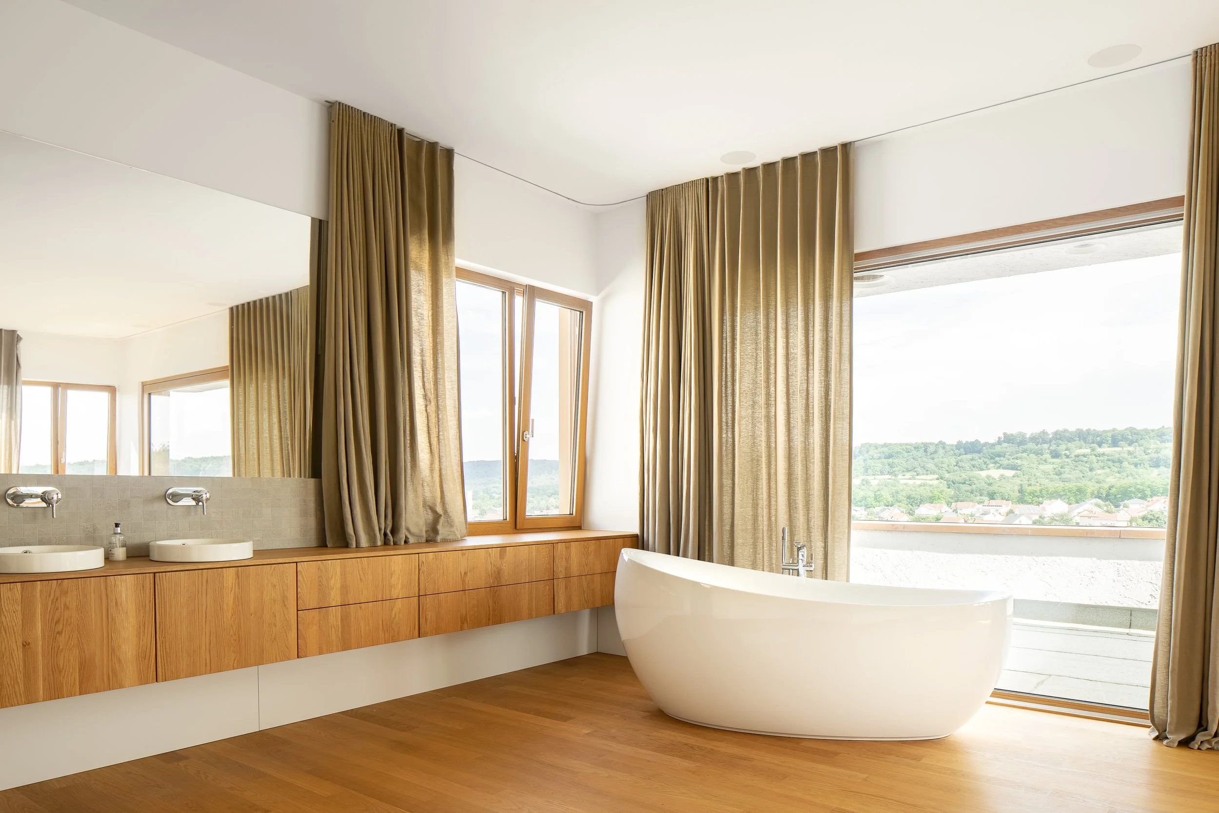 Ein modernes Badezimmer mit einer freistehenden Badewanne, großen Fenstern, goldenen Vorhängen und Holzboden.